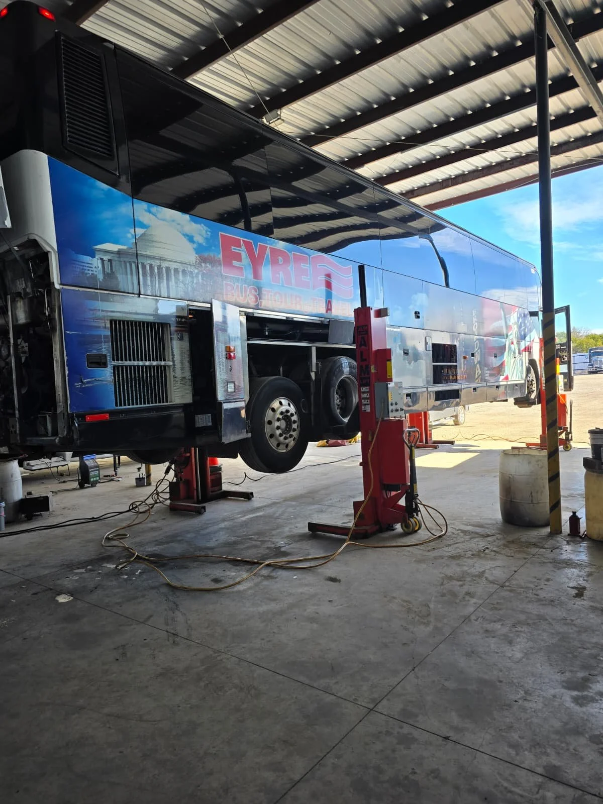 A large sightseeing bus with advertising for Eyre Bus Tour on the side, raised on hydraulic lifts inside a garage or maintenance facility with tools and equipment around.