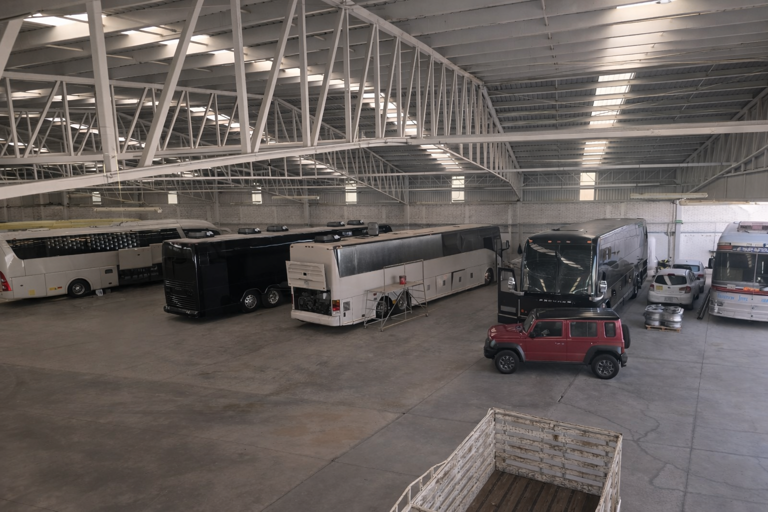 Indoor parking area with multiple buses, vans, and a red Jeep. The space has a high ceiling with white metal beams and large overhead lighting.