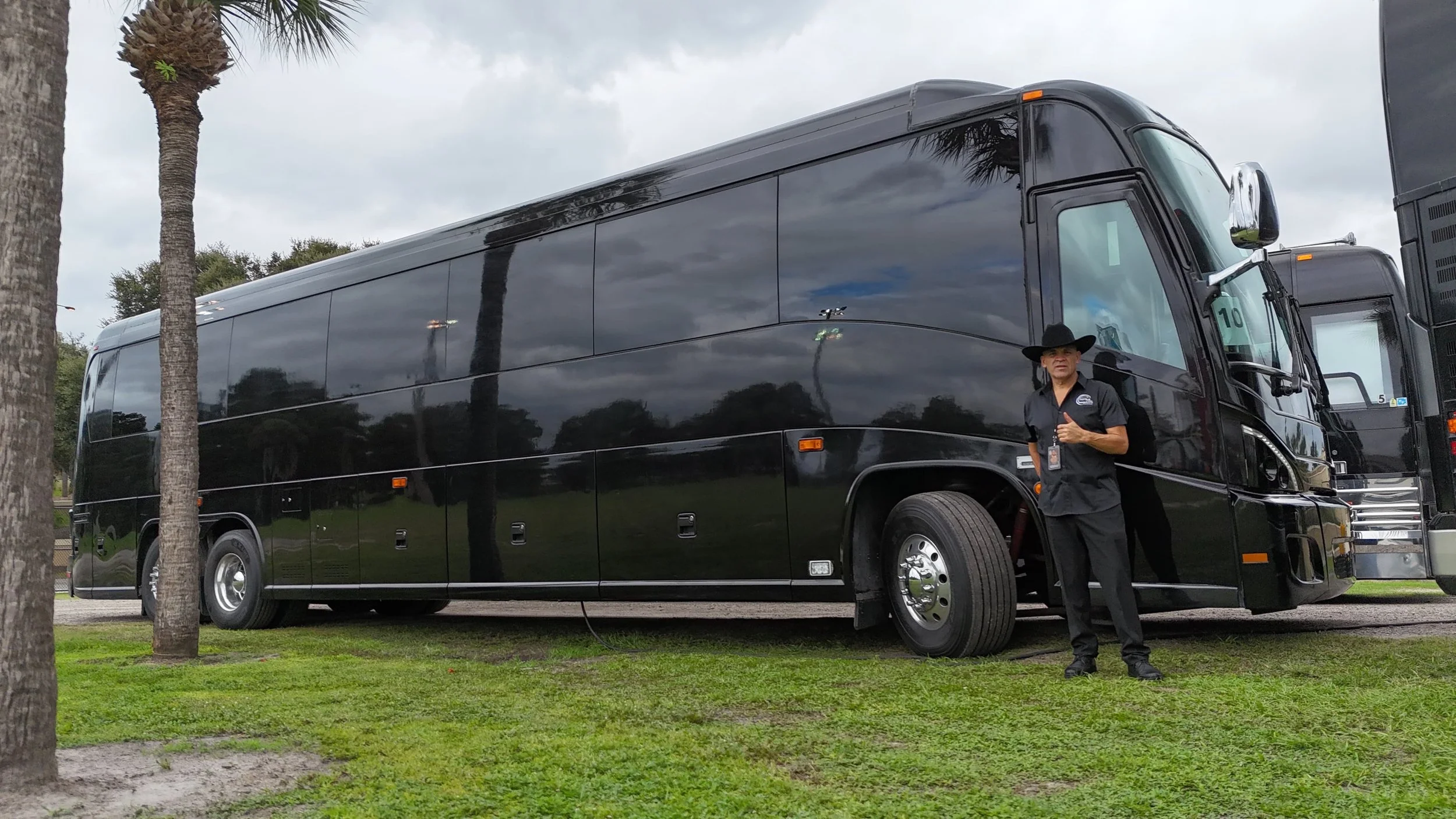 A person dressed in black clothing, including a wide-brimmed hat, stands next to a large black bus on a grassy area with trees and cloudy sky in the background.