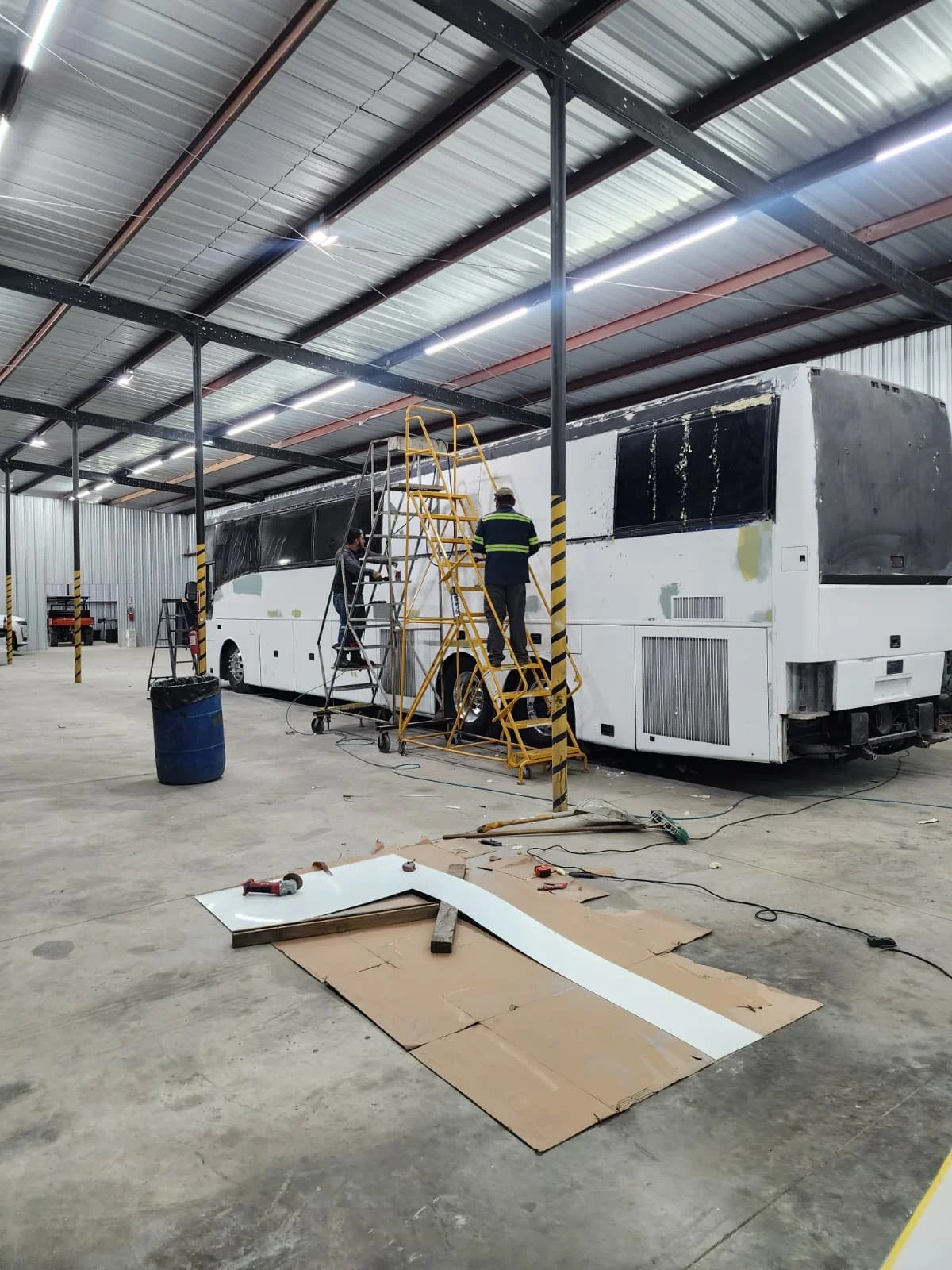 Workers repairing or renovating the exterior of a bus inside a large warehouse or garage.