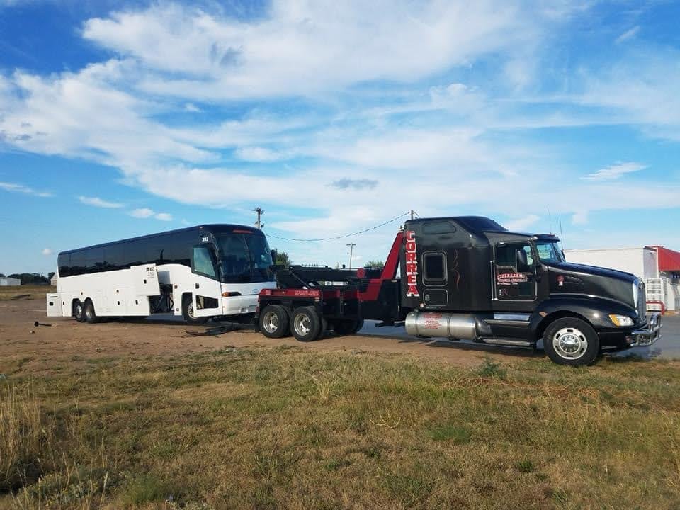 A black semi-truck with the word "Goreea" on its side is towing a large white bus on a rural road, with a blue sky and some clouds in the background.