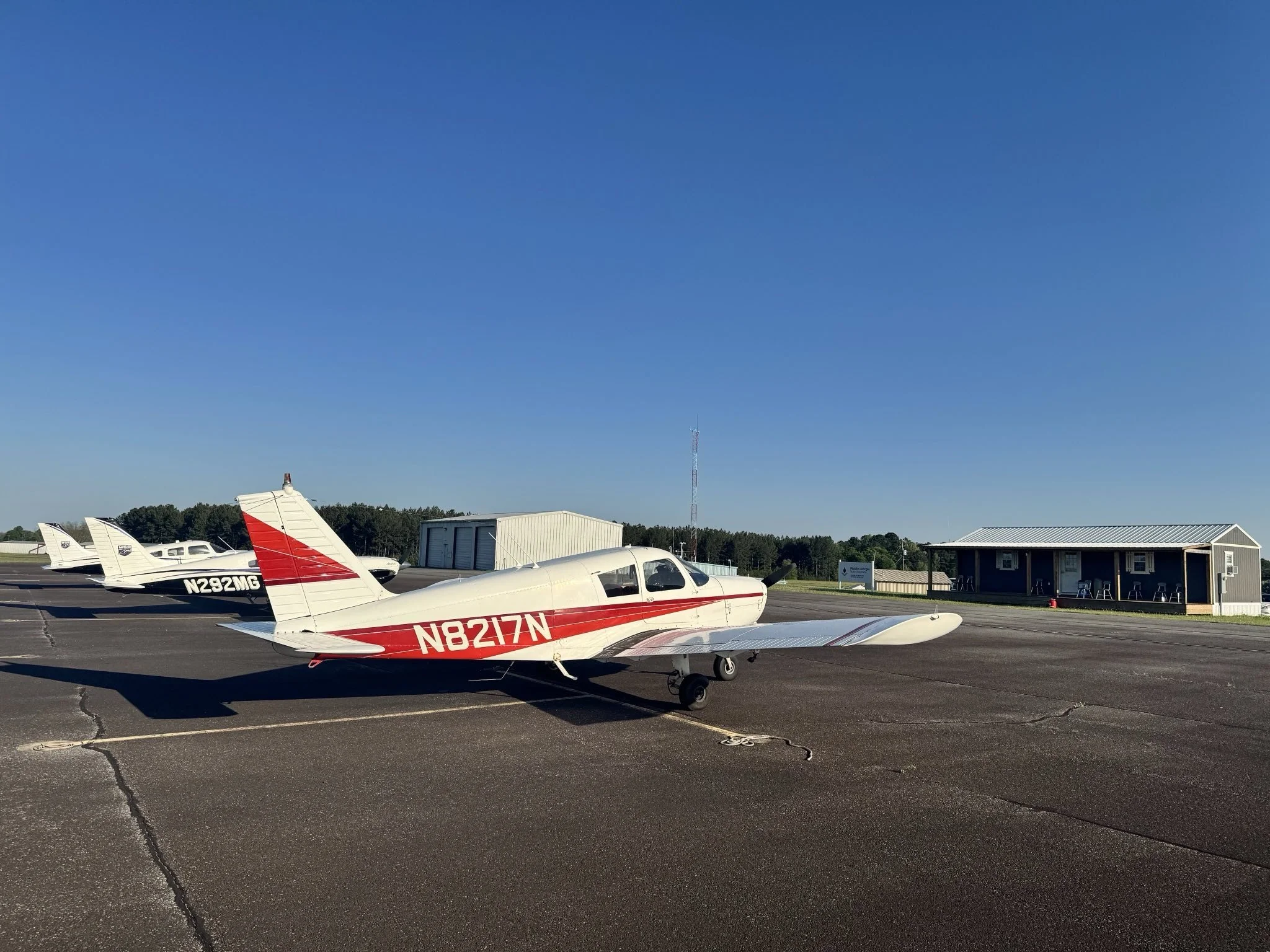 Small airport tarmac with three small propeller planes parked, one prominently in the foreground with red and white markings, and a hangar and building in the background under a clear blue sky.