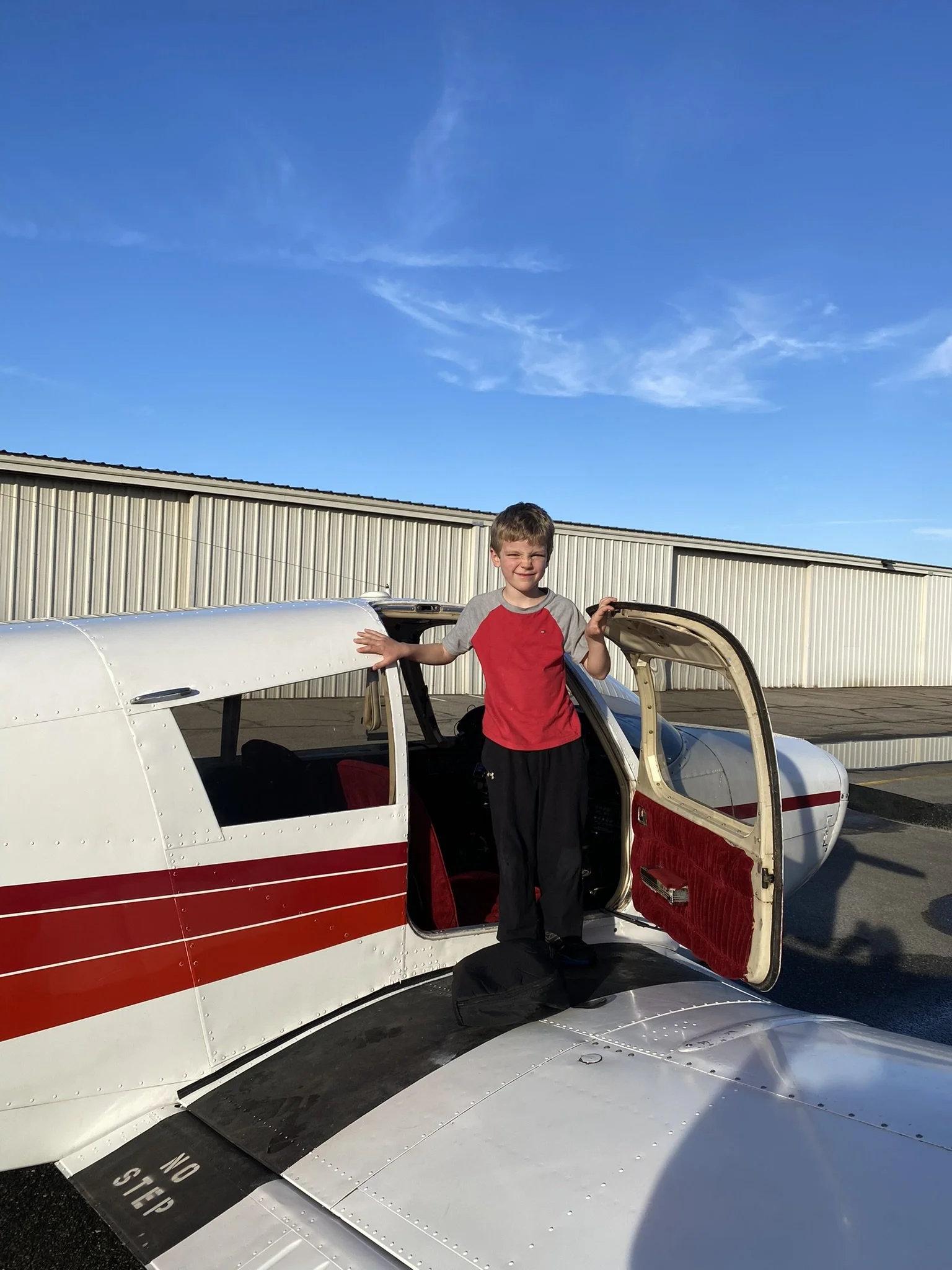 A young boy standing inside an airplane with the cockpit door open, at an airport tarmac, under a clear blue sky.