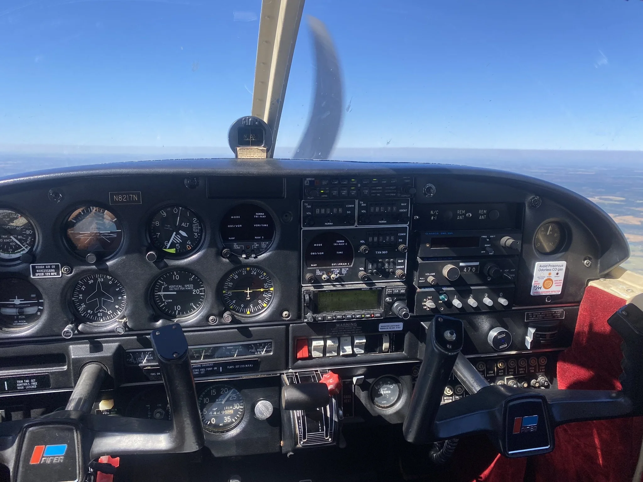 The cockpit of a small airplane with various gauges, switches, and controls, flying high above a landscape with a clear blue sky.