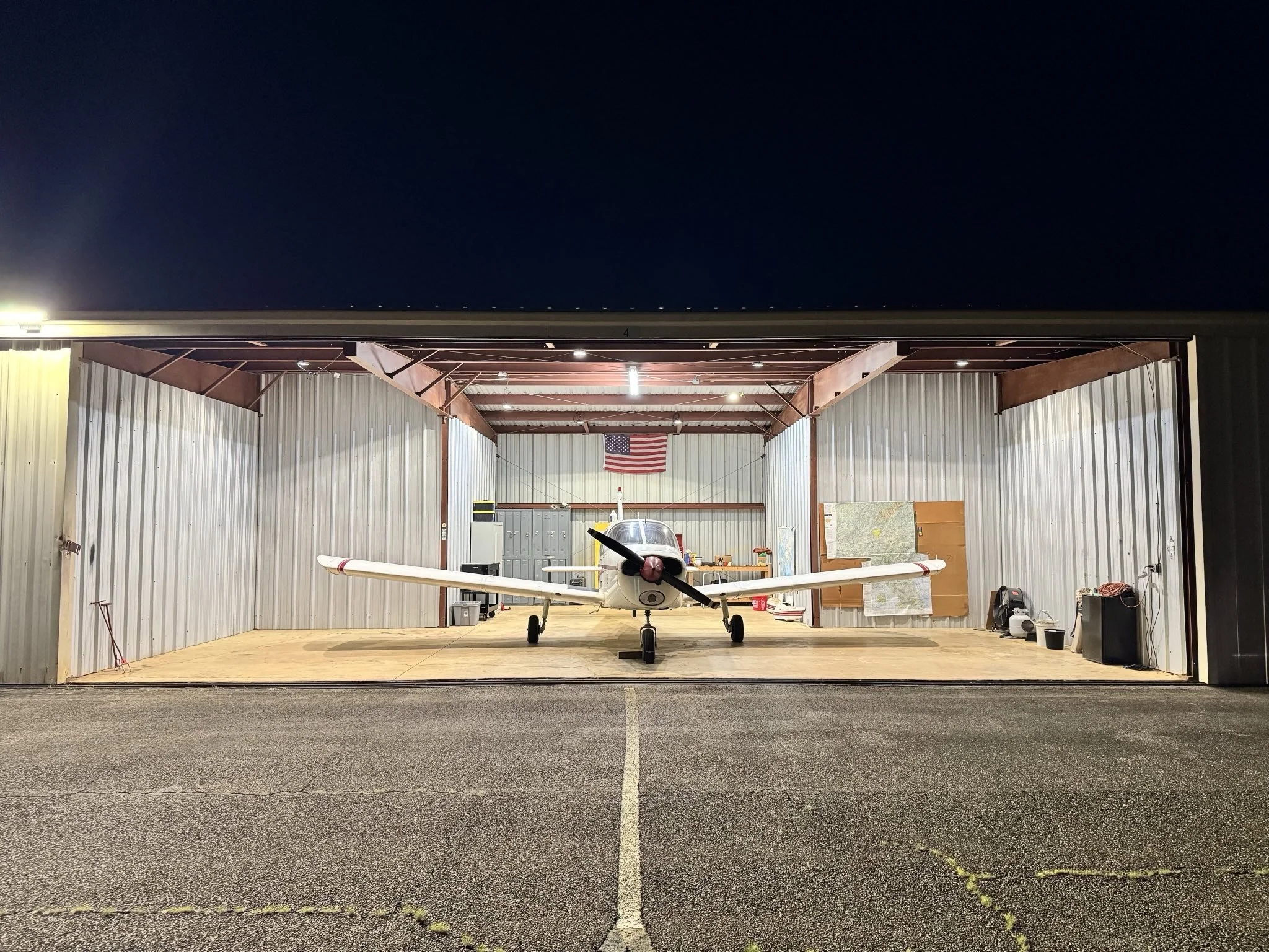 Small aircraft parked inside a metal airplane hangar at night, with an American flag hanging on the back wall.