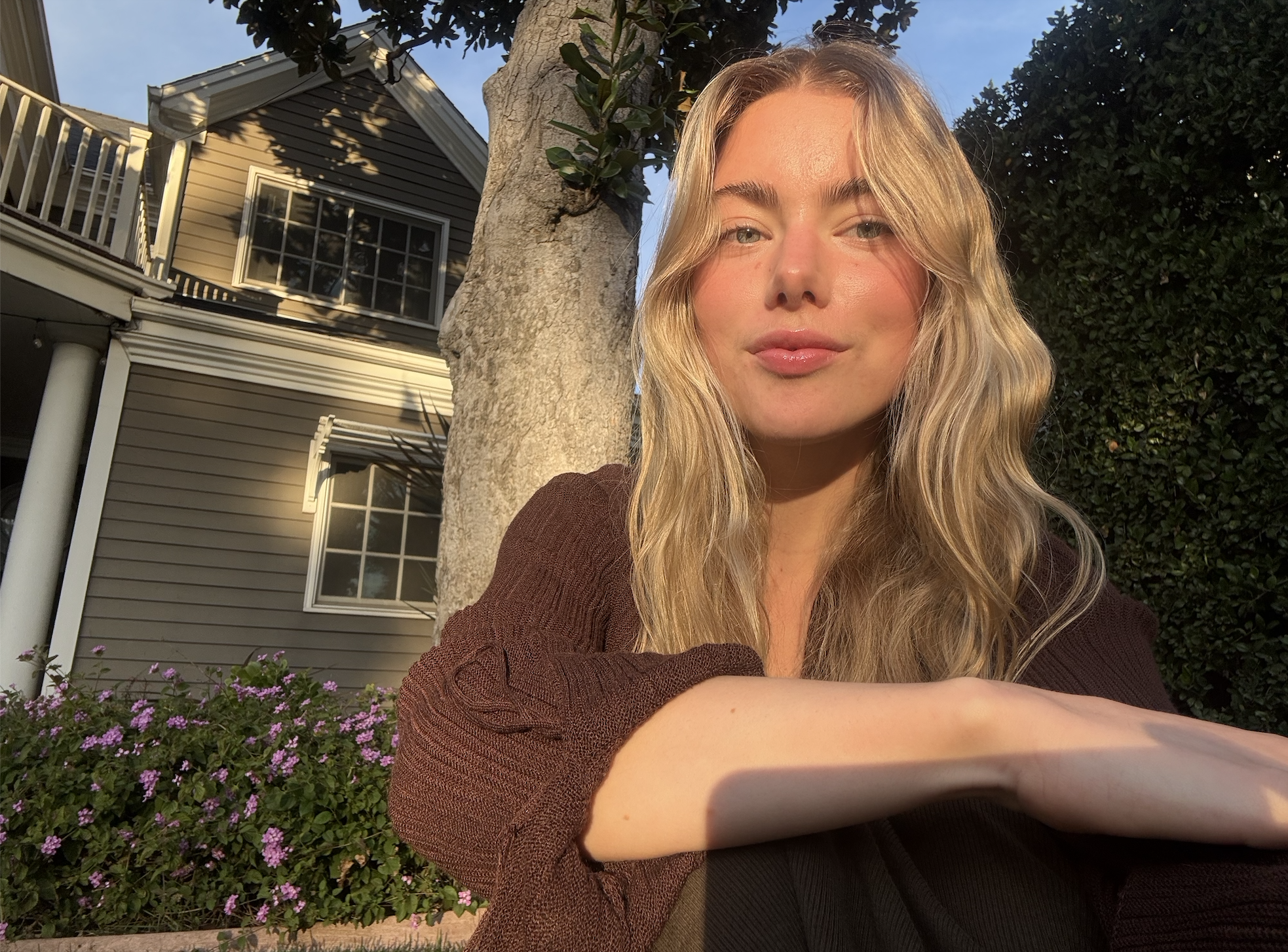 A young woman with blonde wavy hair smiling in sunlight during golden hour, leaning on her arm in front of a house with purple flowers and a large tree.