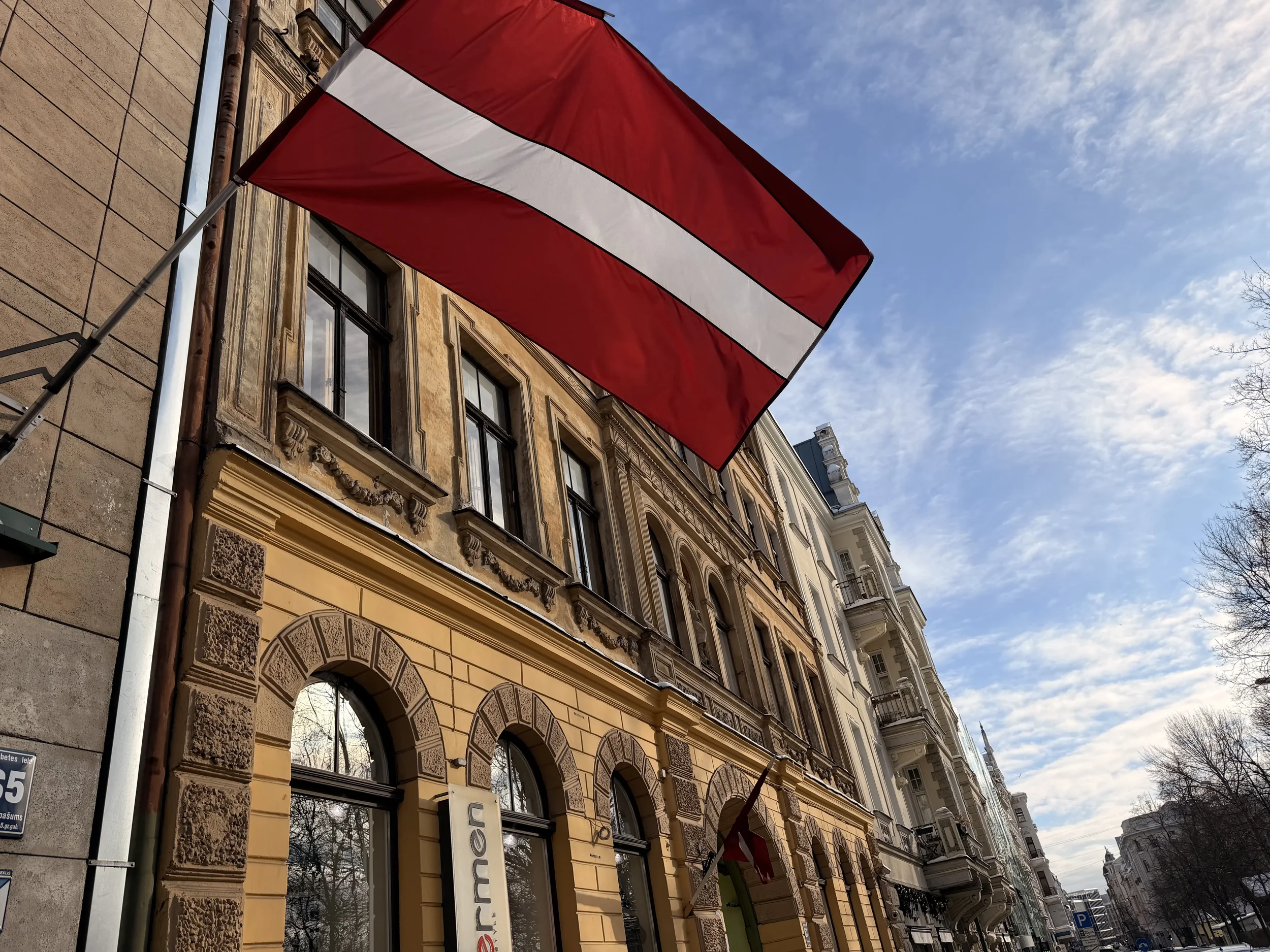 Latvian flag waving above historic buildings along a street in Riga under a clear sky.