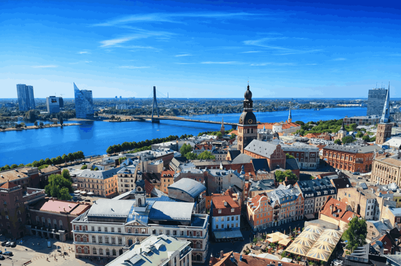 Aerial view of Riga, Latvia, showing the historic Old Town with colorful rooftops, church spires, and the Daugava River under a clear blue sky.