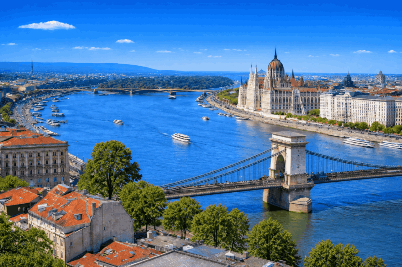 Daytime aerial view of Budapest, Hungary, featuring the Danube River, Chain Bridge, and the Hungarian Parliament Building under a clear blue sky.