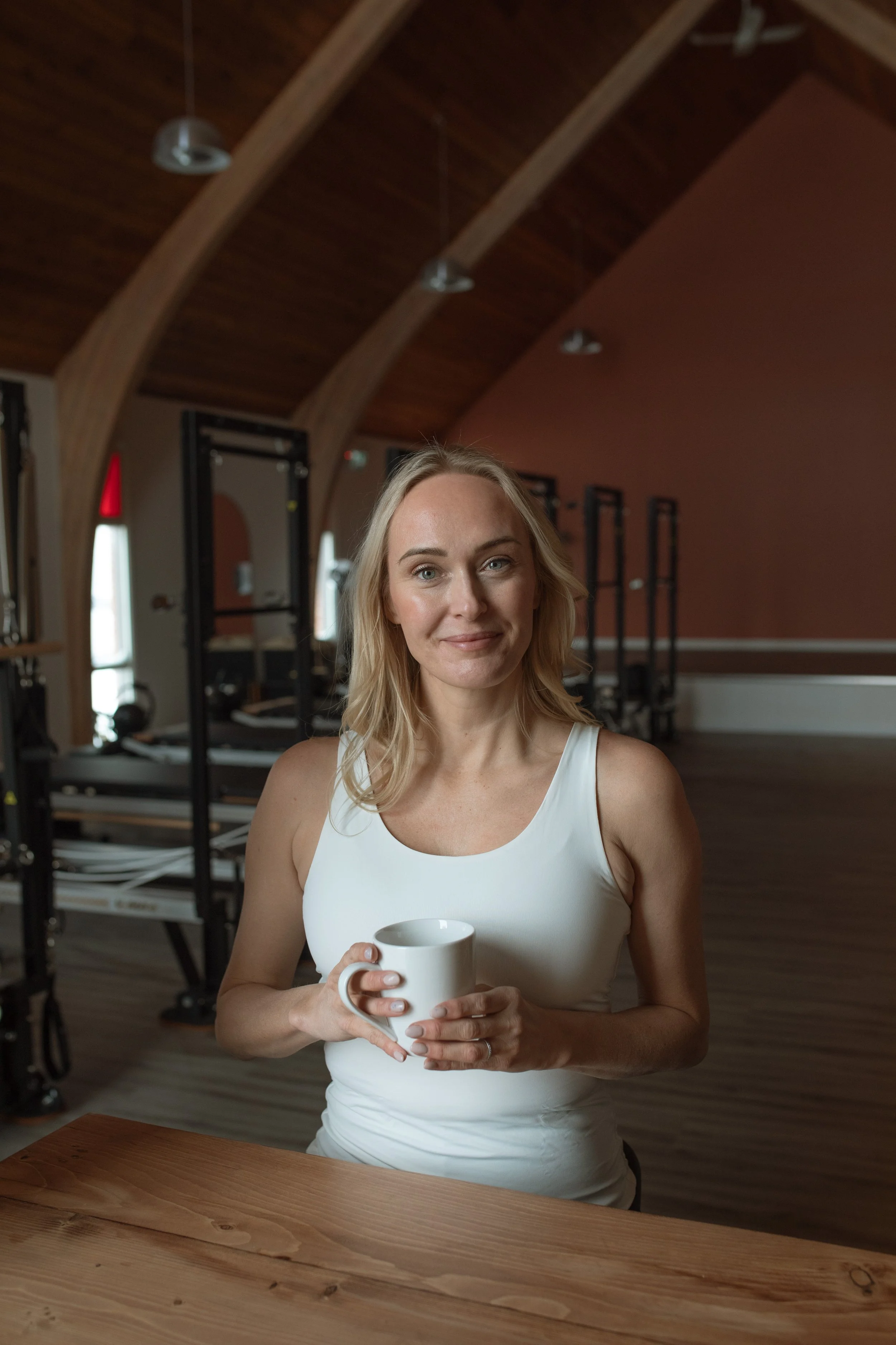 A woman with blonde hair wearing a white tank top, holding a white mug, and smiling while sitting at a wooden table in a gym or fitness studio.