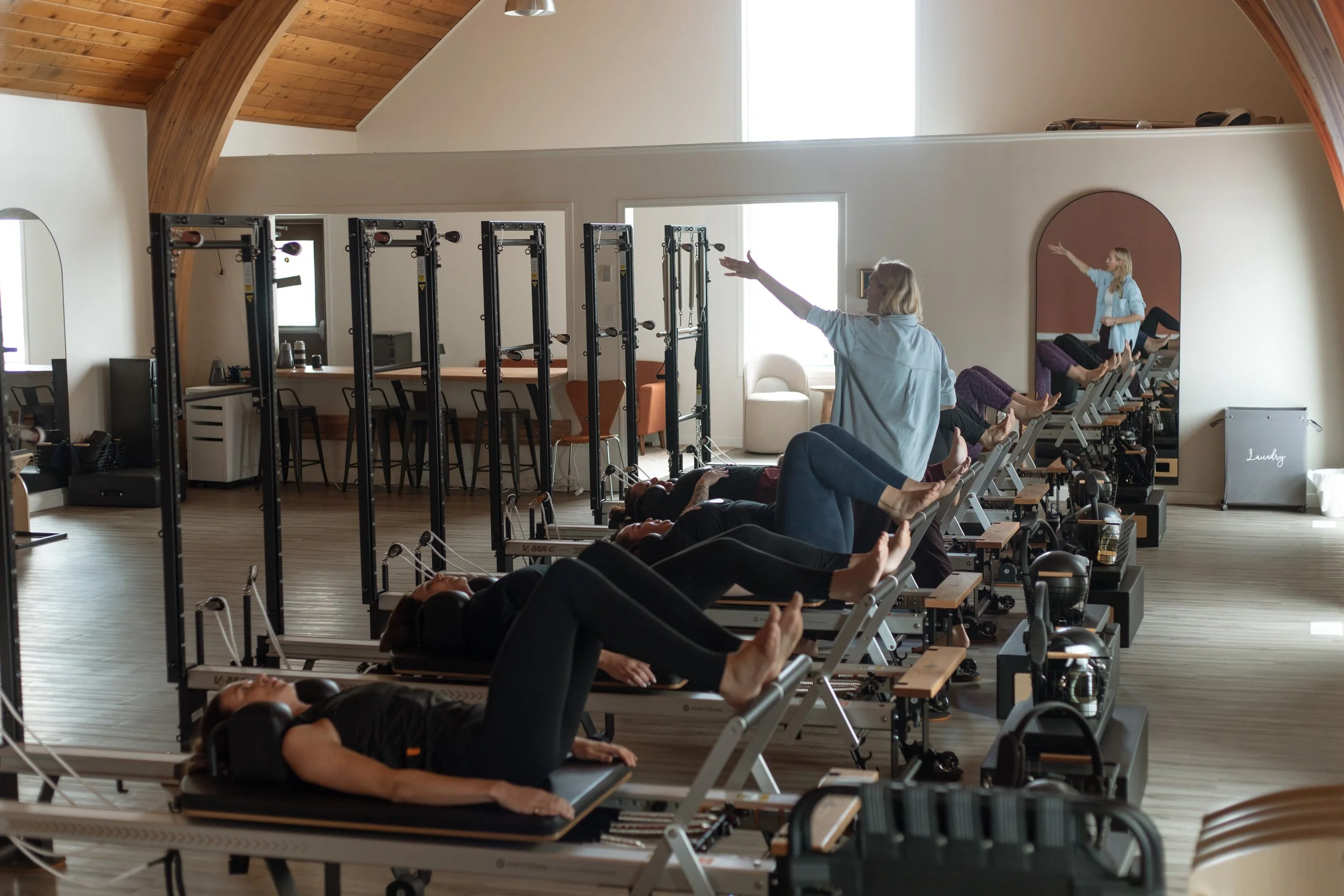 A group exercise class in a pilates studio with people lying on reformer machines, instructor at the front giving instructions, mirror on the wall, and natural light coming through windows.