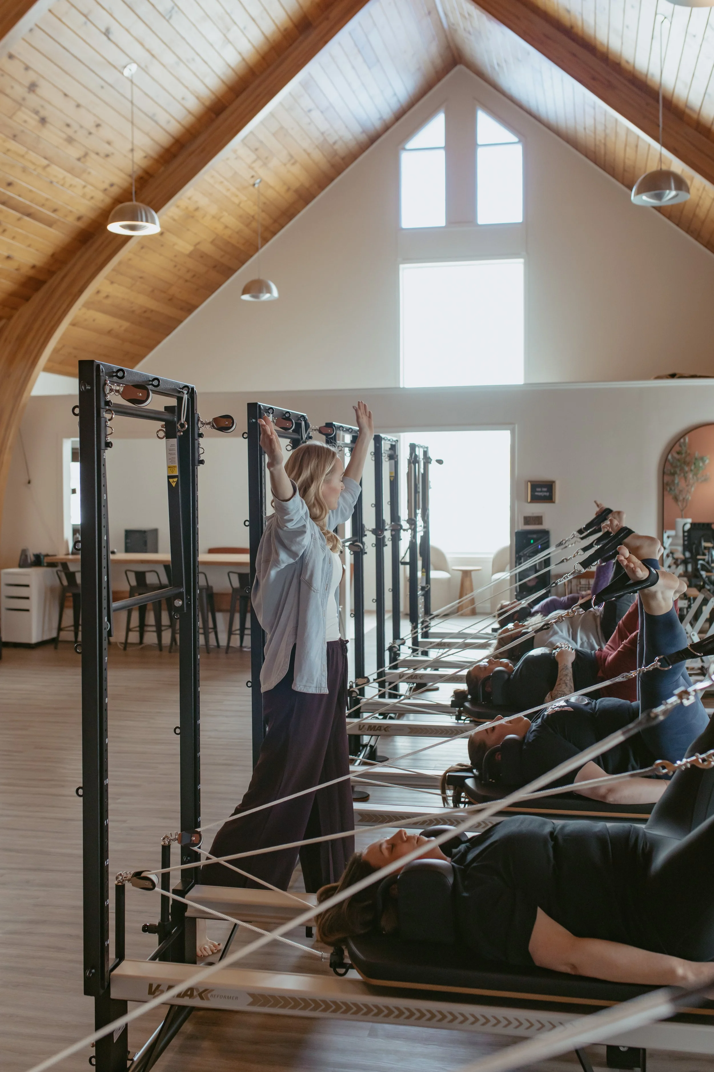 A fitness instructor leading a pilates class on reformer machines in a spacious, well-lit gym with wooden ceiling and high windows.