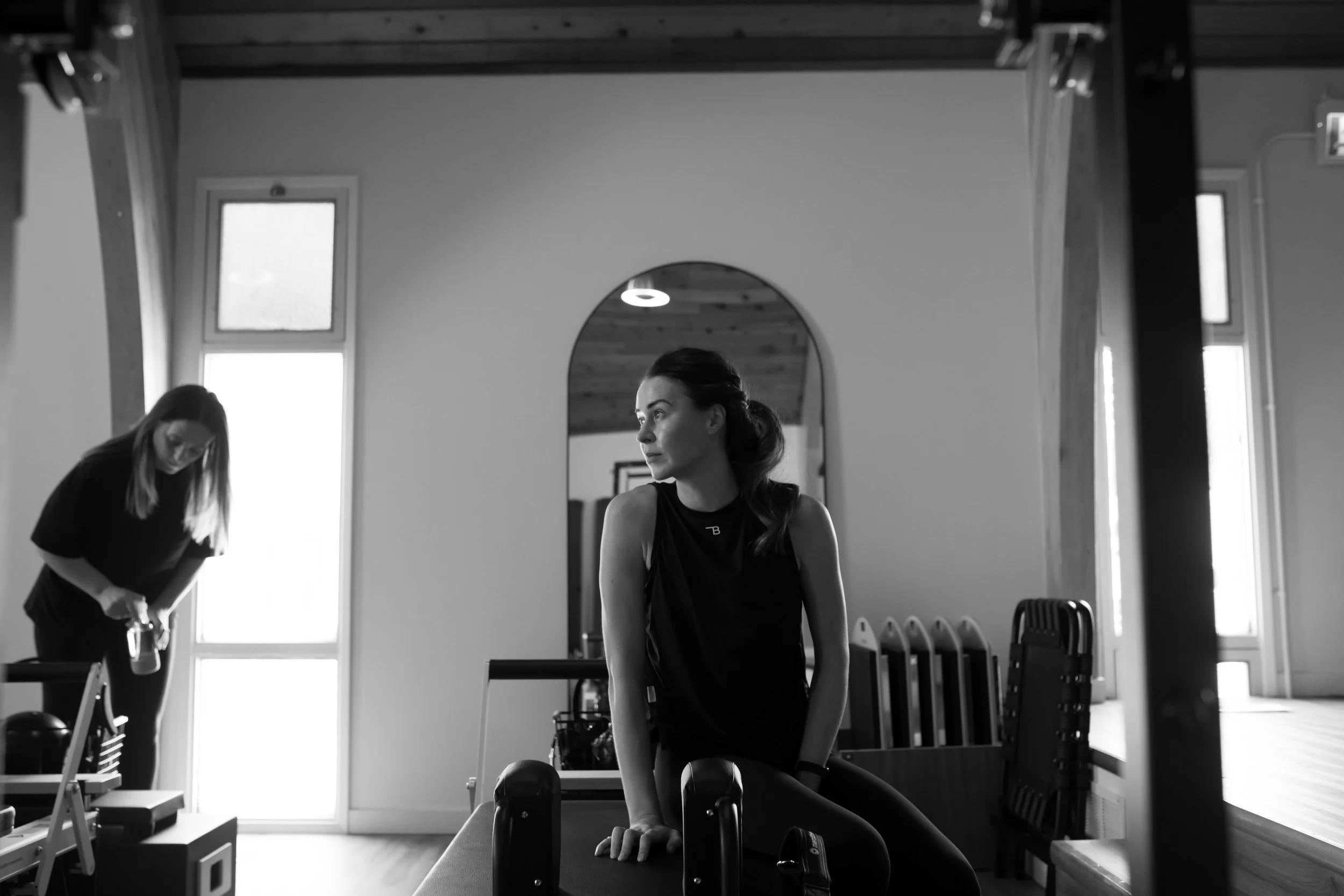A woman sitting on a bench in a modern gym, looking to the side, with a trainer in the background preparing equipment, all in black and white.