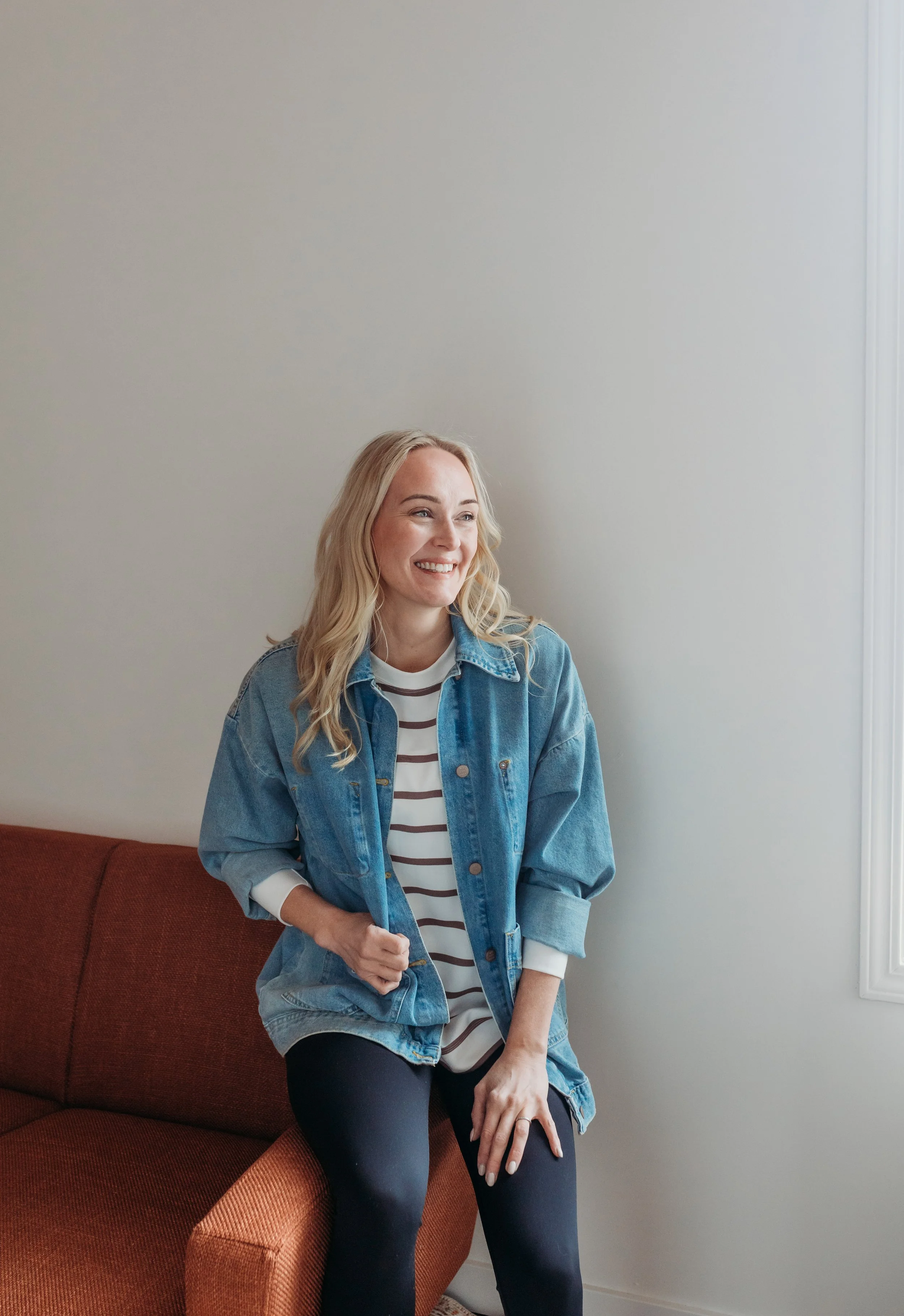 A young woman with blonde hair sitting on a red sofa, smiling and looking to her right, wearing a denim jacket over a striped shirt and black pants, in a room with white walls and a window.