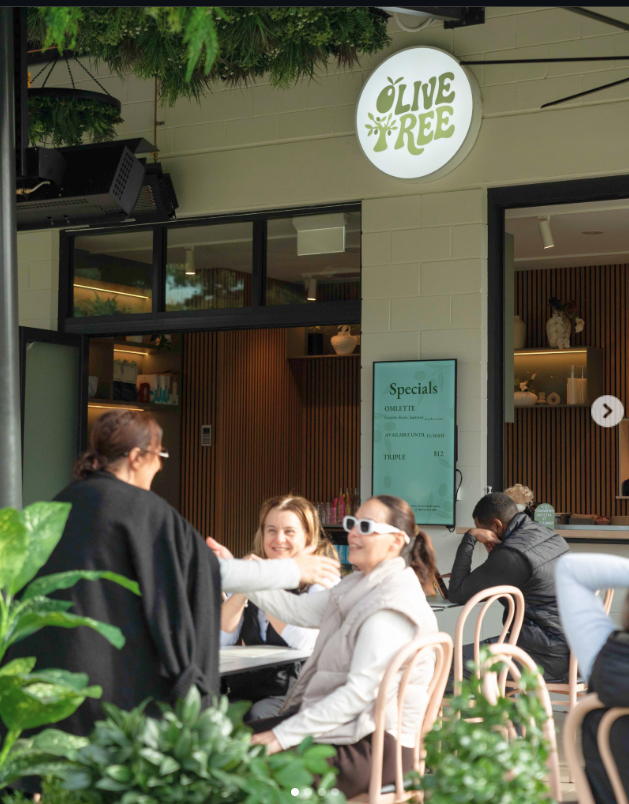 People sitting at an outdoor café, smiling and talking, with a sign that says 'Olive Tree' above the entrance.