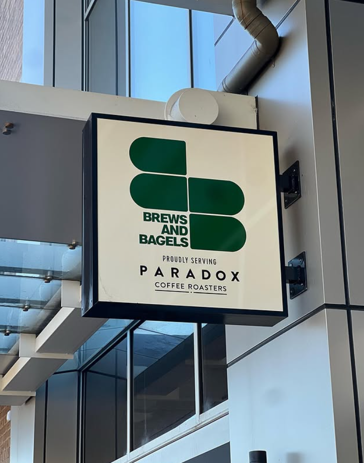 A square sign with a large green and black logo and text reading 'Brews and Bagels'. Additional text below says 'Proudly Serving Paradox Coffee Roasters'. The sign is mounted on a building exterior near a glass window and door, with a ventilation pipe above.