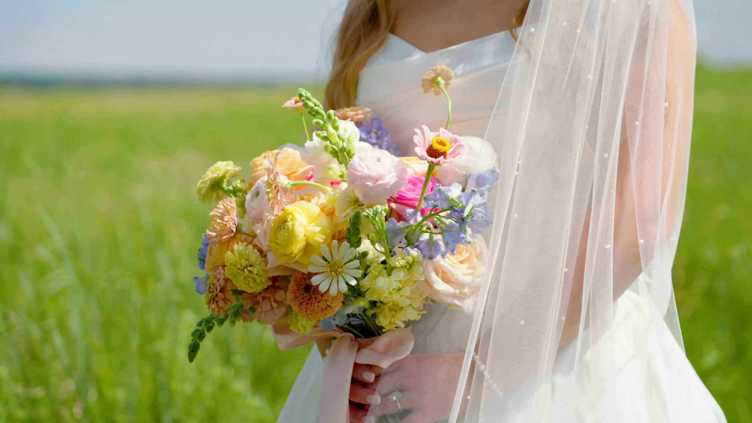 A bride holding a colorful bouquet of flowers outdoors in a green field, wearing a white wedding dress and veil.