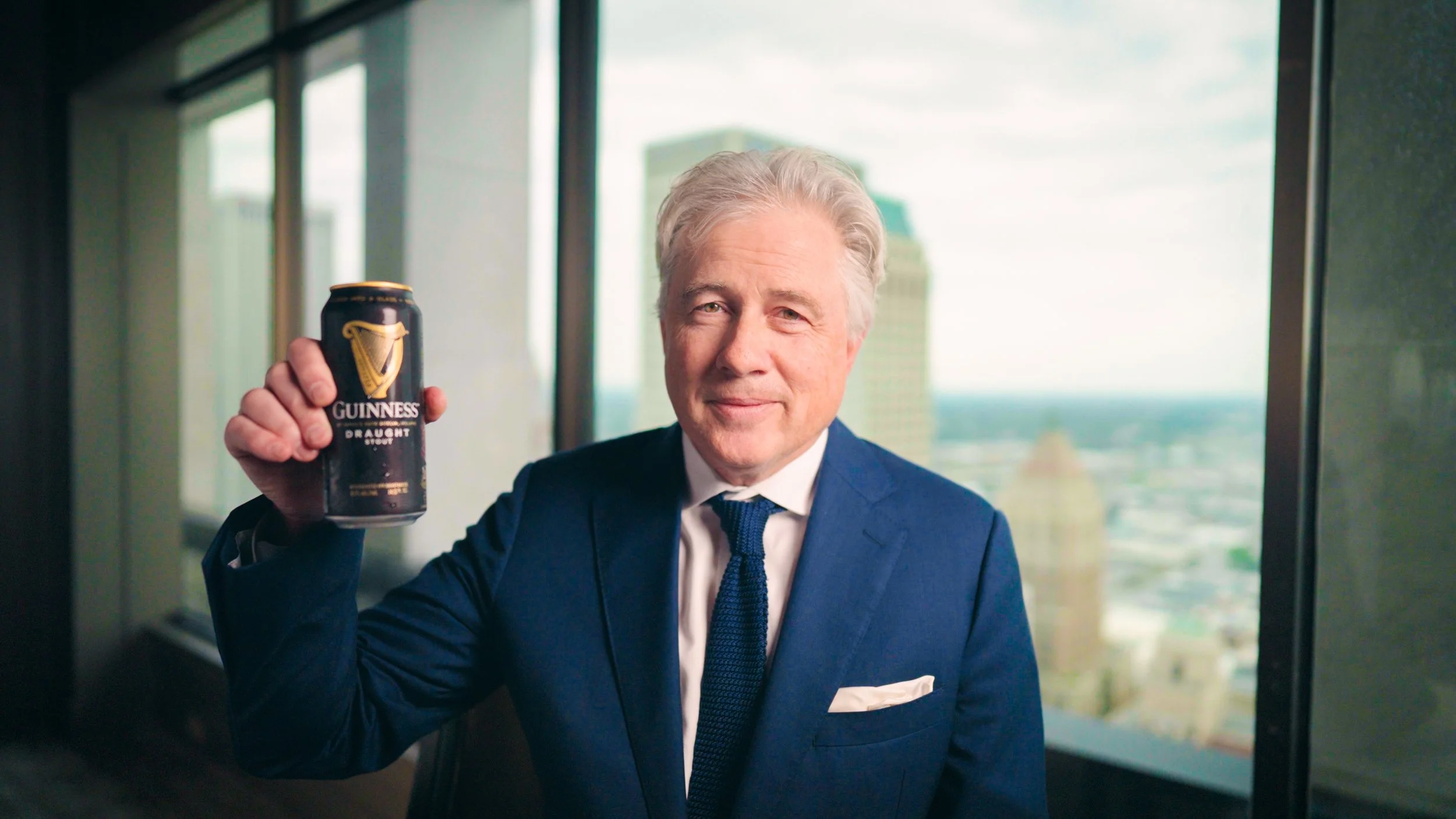 A man in a blue suit and tie holding a can of Guinness beer, standing in front of a large window with a city view.