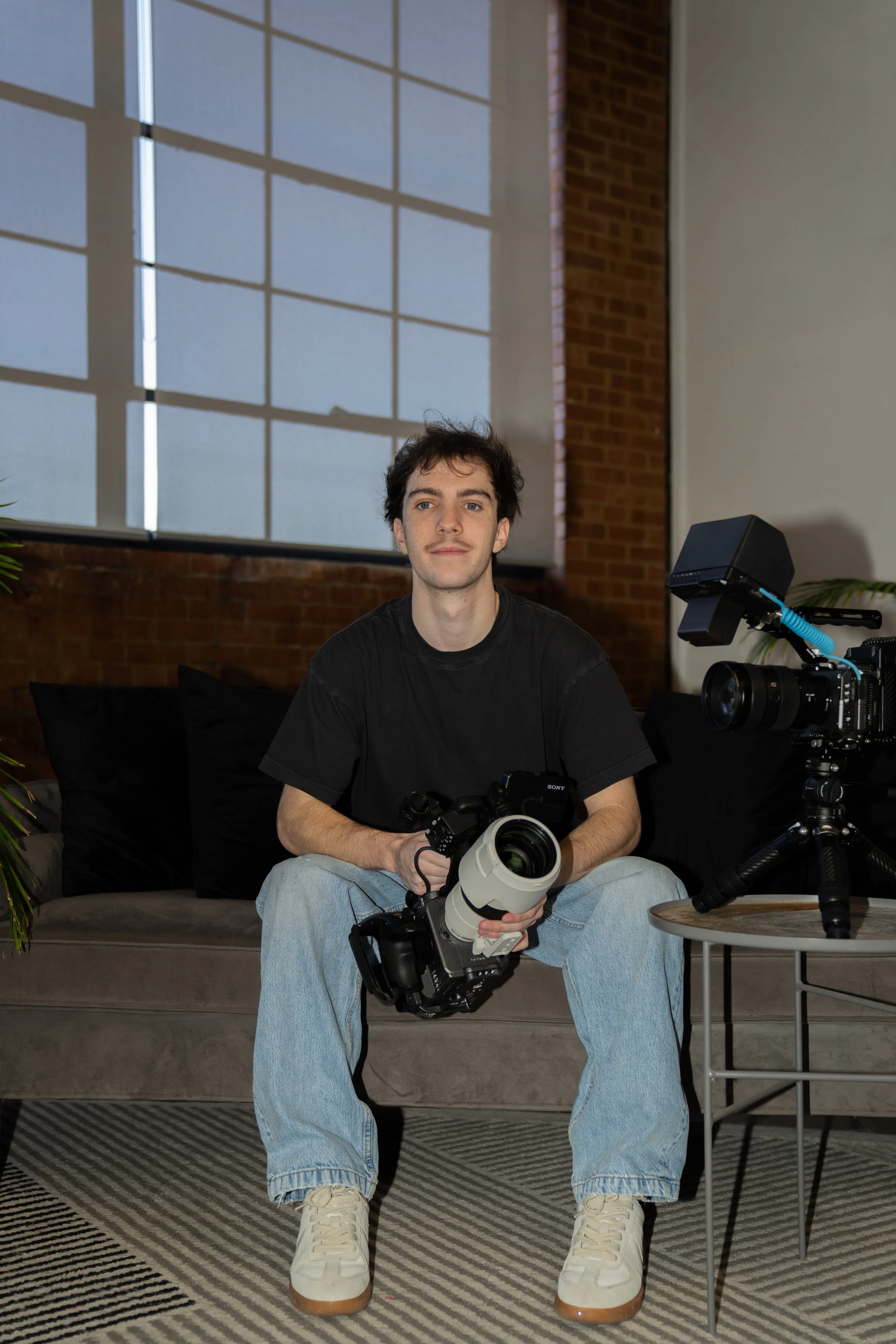 A young man sitting on a gray sofa holding a professional camera with a large telephoto lens in a studio or office setting.