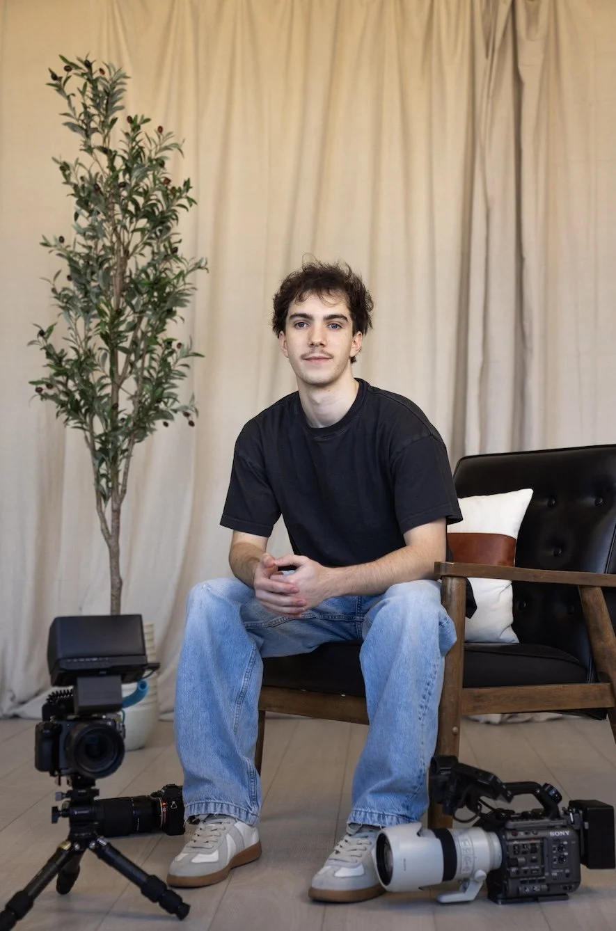 A young man sitting on a chair with cameras on the floor in front of him, in a room with beige curtains and a potted plant in the background.
