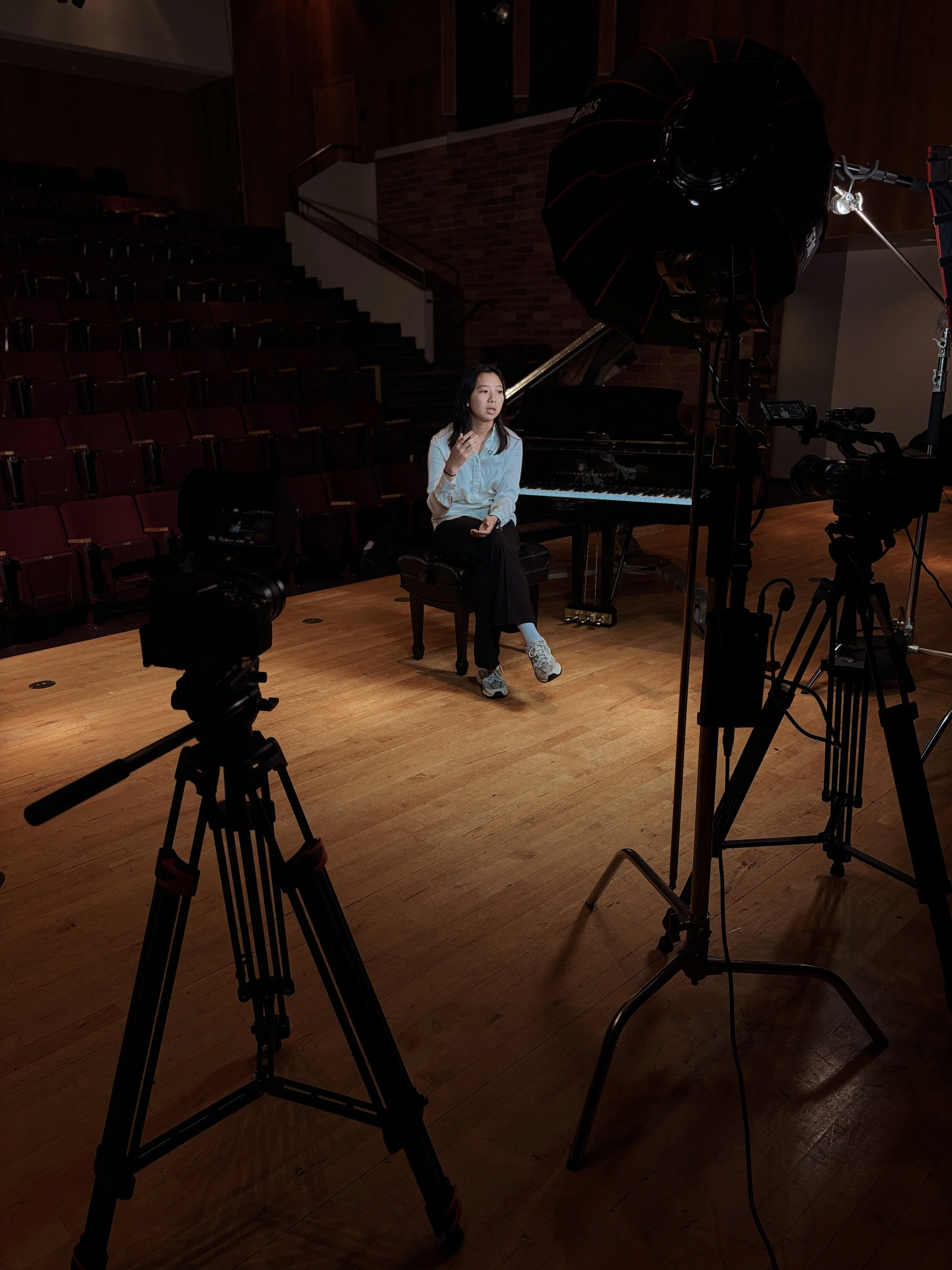A woman sitting on a stool in a music rehearsal room, surrounded by professional filming and lighting equipment, with a grand piano in the background.