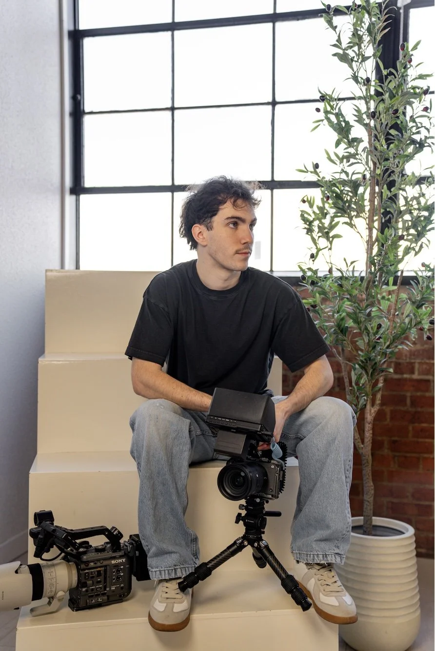 A young man with dark curly hair and light skin sitting on a cream-colored bench indoors, holding a camera on a tripod in front of him. There is a video camera on the ground to his left, and a potted plant with dark berries to his right, against a background of large windows and brick wall.
