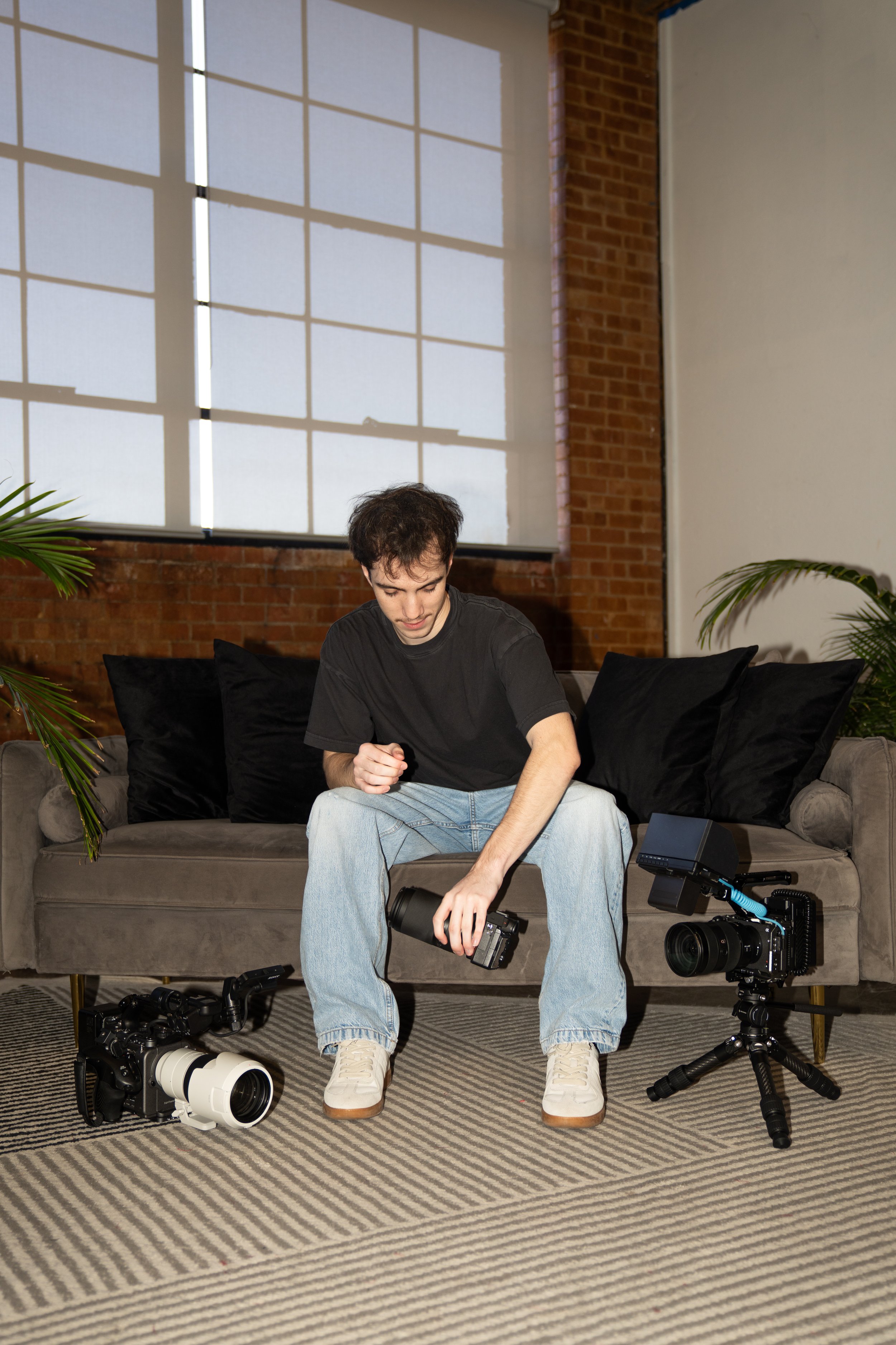 A young man in a black t-shirt and light blue jeans sits on a gray sofa in an industrial-style room with brick walls and large windows, surrounded by professional camera equipment, including two cameras and a tripod, arranging or inspecting a camera accessory.