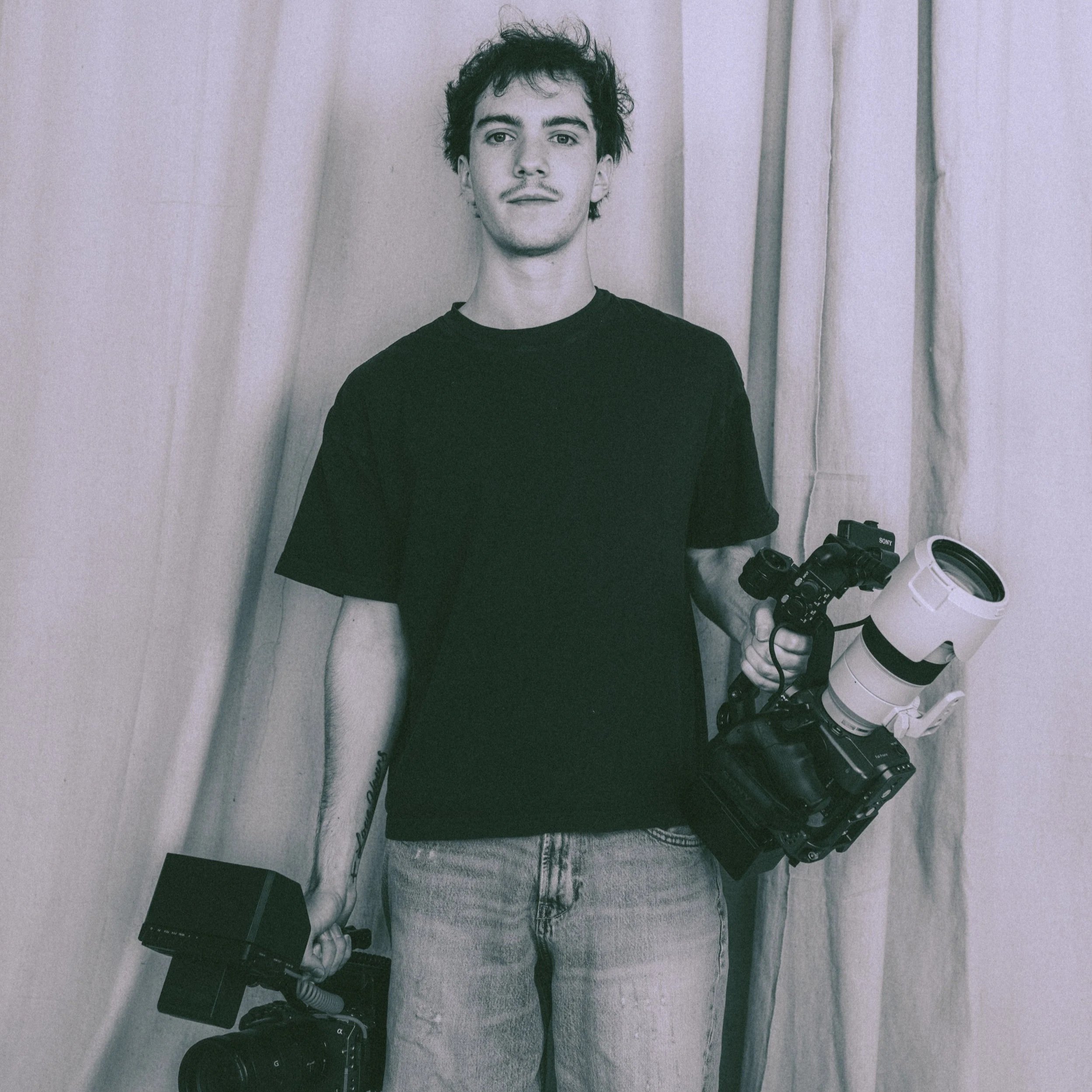 A young man with dark curly hair, wearing a black T-shirt and jeans, holding a camera in each hand, standing in front of a curtain.