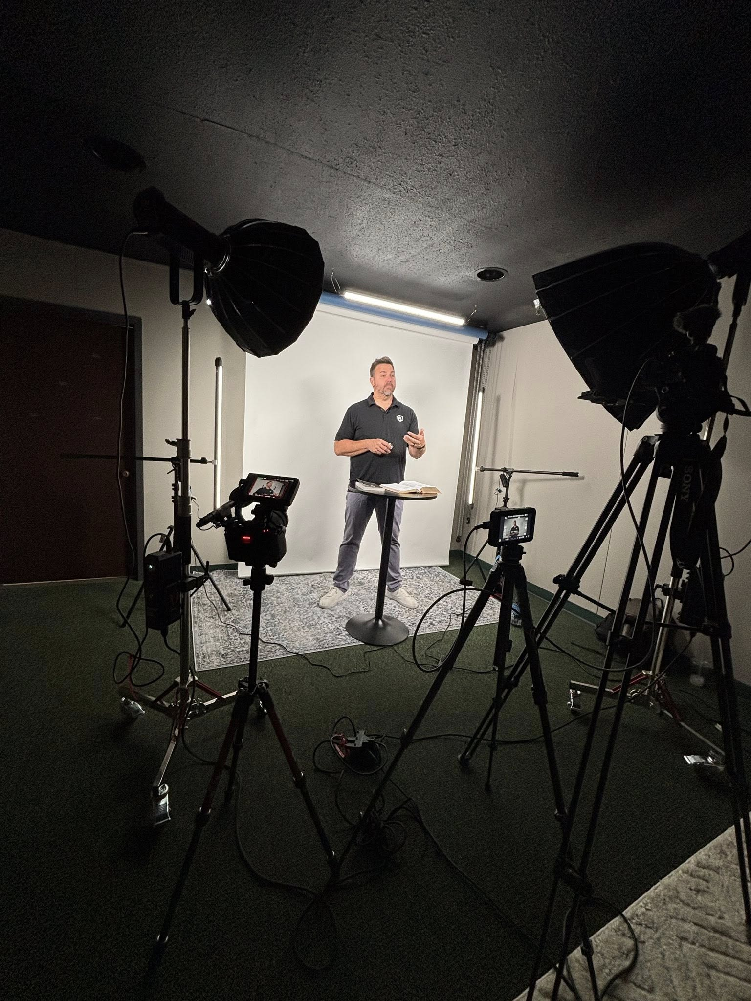 A man is standing in front of a white backdrop, speaking or presenting, with a book or notes on a small table in front of him. The scene is set up with professional lighting and cameras, indicating a recording or live streaming session.