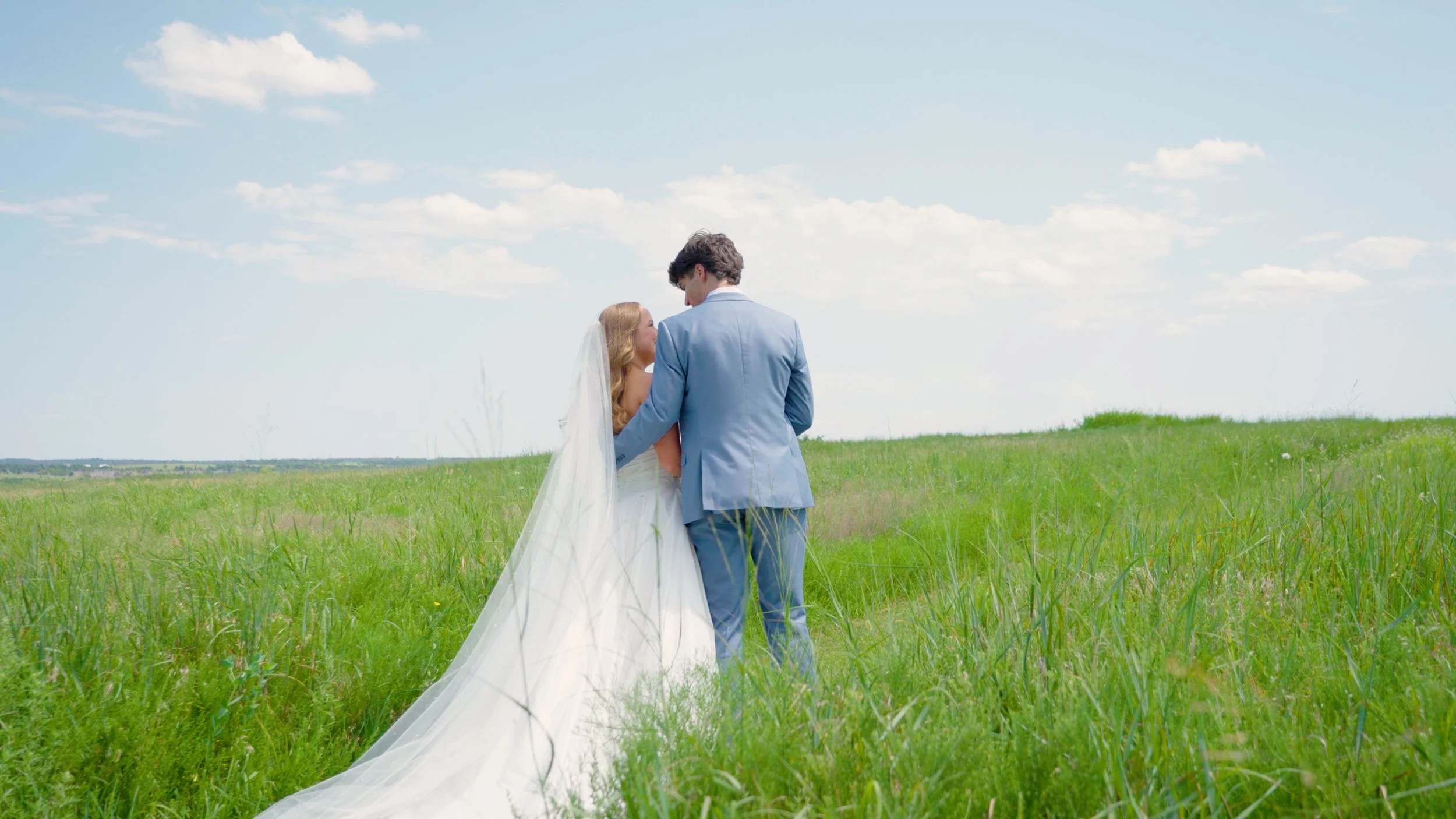 A bride and groom standing in a grassy field under a partly cloudy sky, embracing each other during their wedding photo shoot.