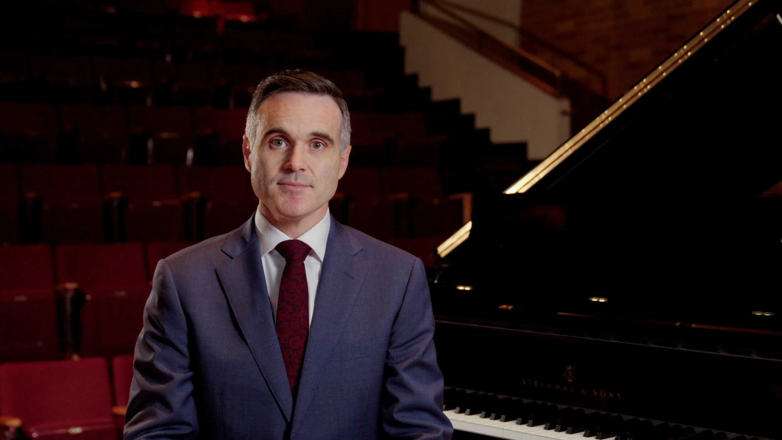 A man in a suit standing in front of a grand piano in an auditorium or recital hall