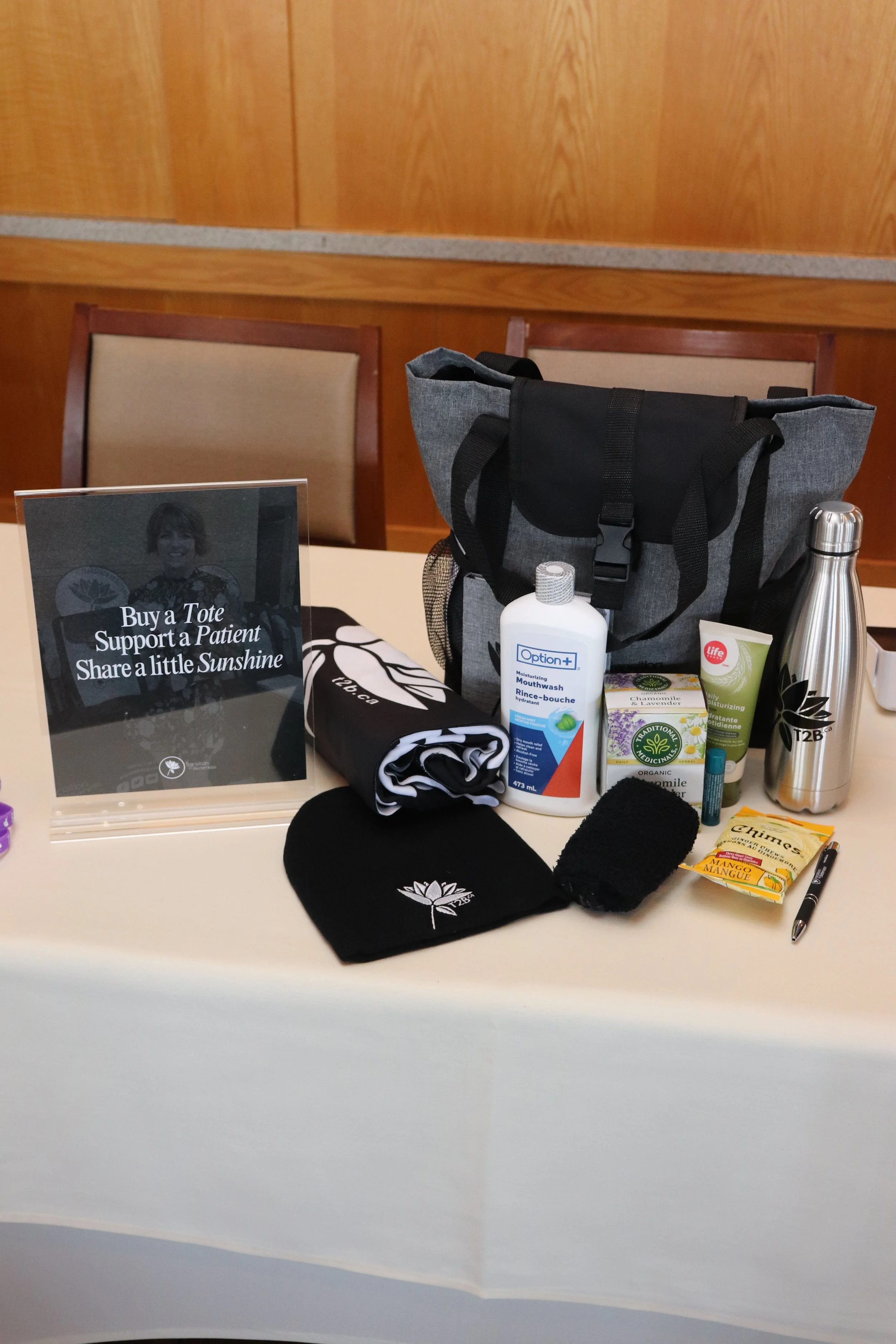 Table with various health and wellness items including a water bottle, mouthwash, herbal products, snacks, a black beanie, an athletic shirt, a tote bag, and a sign encouraging support for patients by buying a tote.