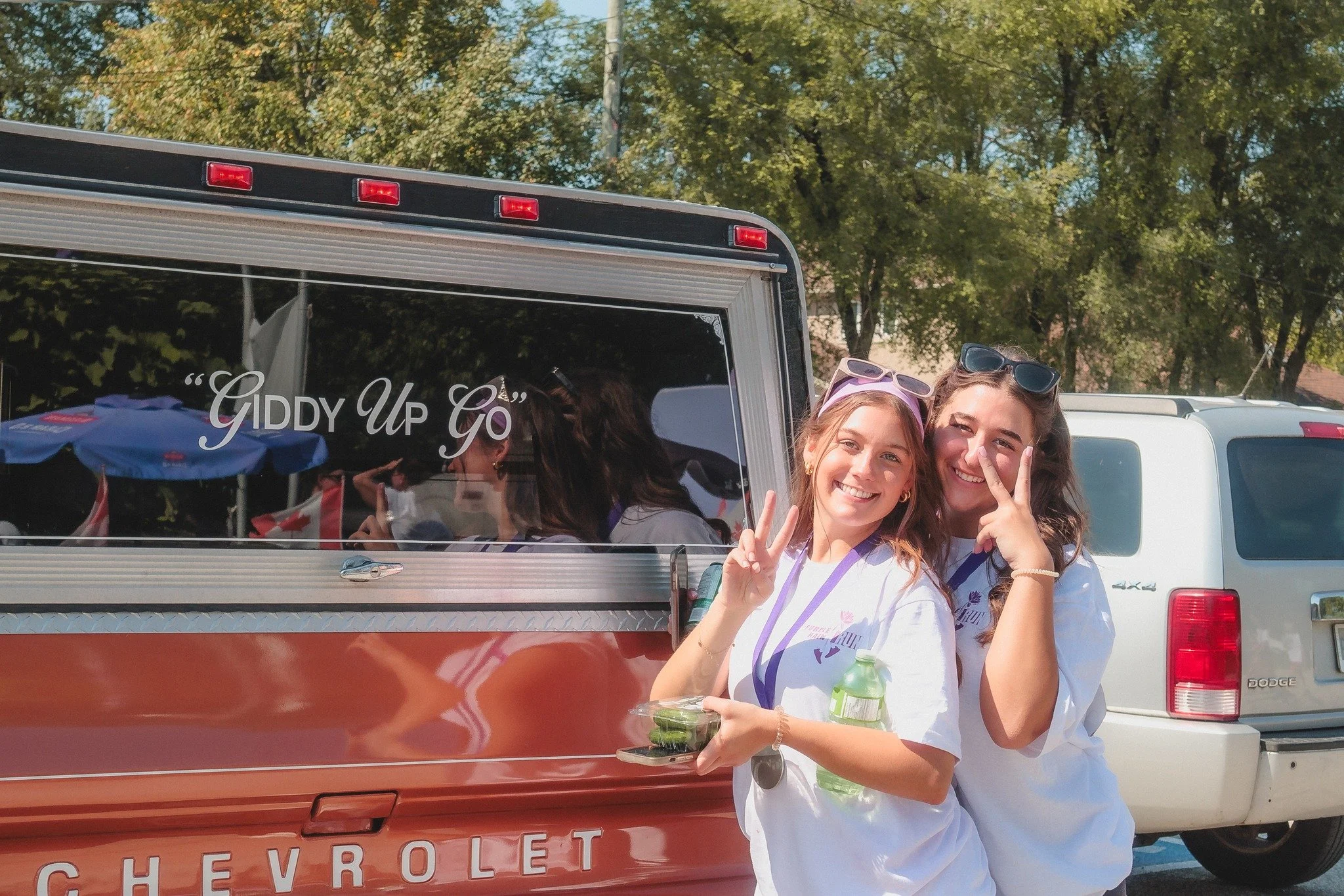 Two young women smiling and making peace signs while standing in front of a vehicle that says "Giddy Up Go" on the window, outdoors with trees in the background.