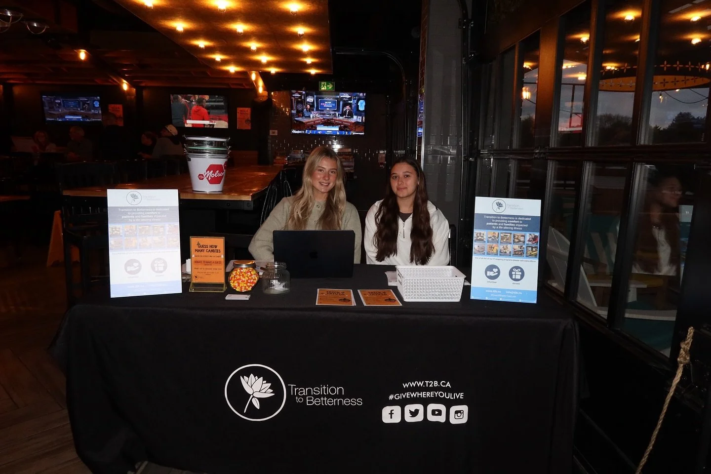 Two young women sitting behind a table with a black tablecloth at an indoor event, promoting Transition to Betterness; one woman has long blonde hair, the other has long dark hair; the table displays informational posters, brochures, a jar of candies, and a large Moesa coffee cup.