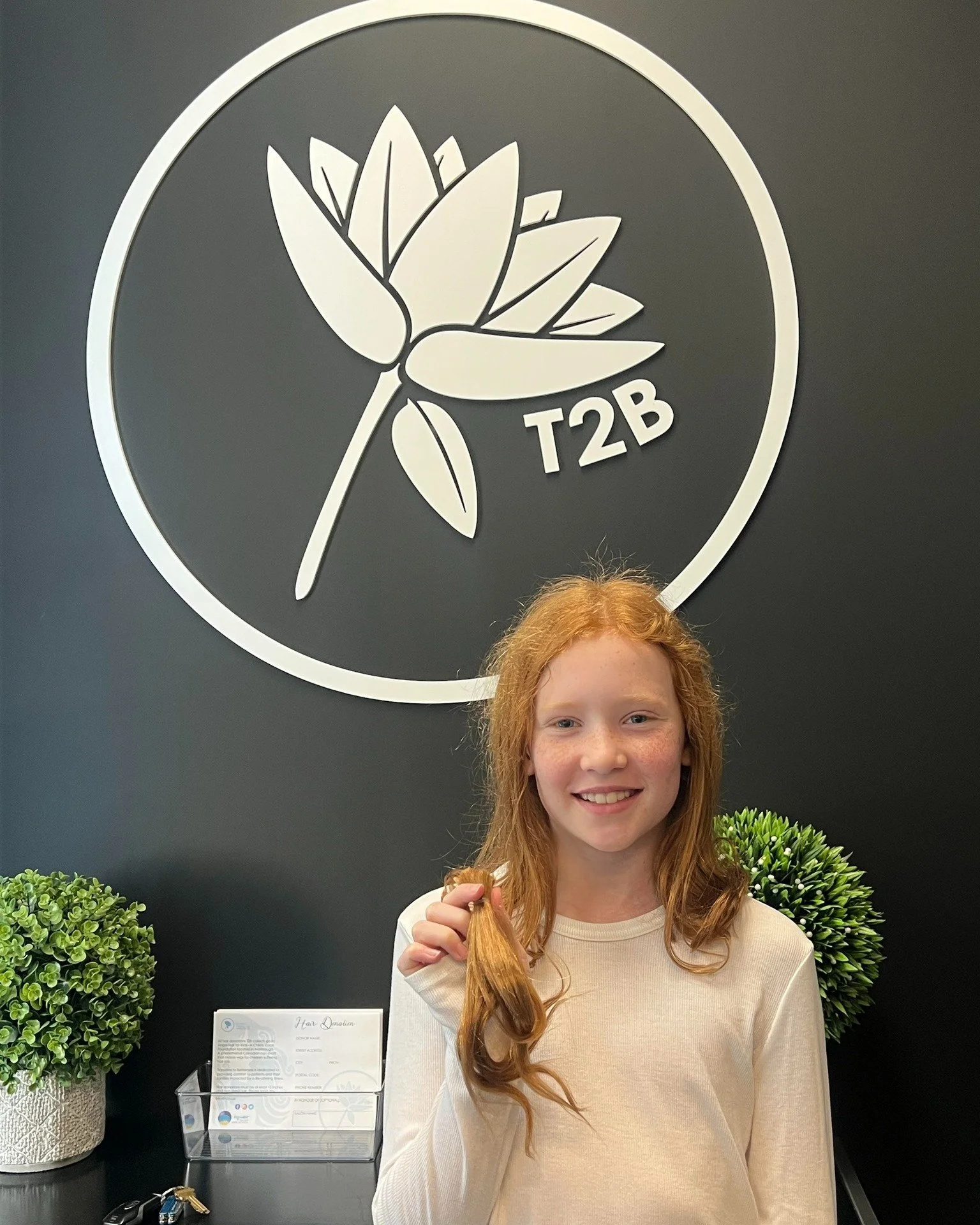 A young girl with red hair and a white shirt holding a lock of her hair, smiling in front of a dark wall with a large logo featuring a stylized flower and the text 'T2B'. Two small potted plants and a business card holder are on the desk beside her.