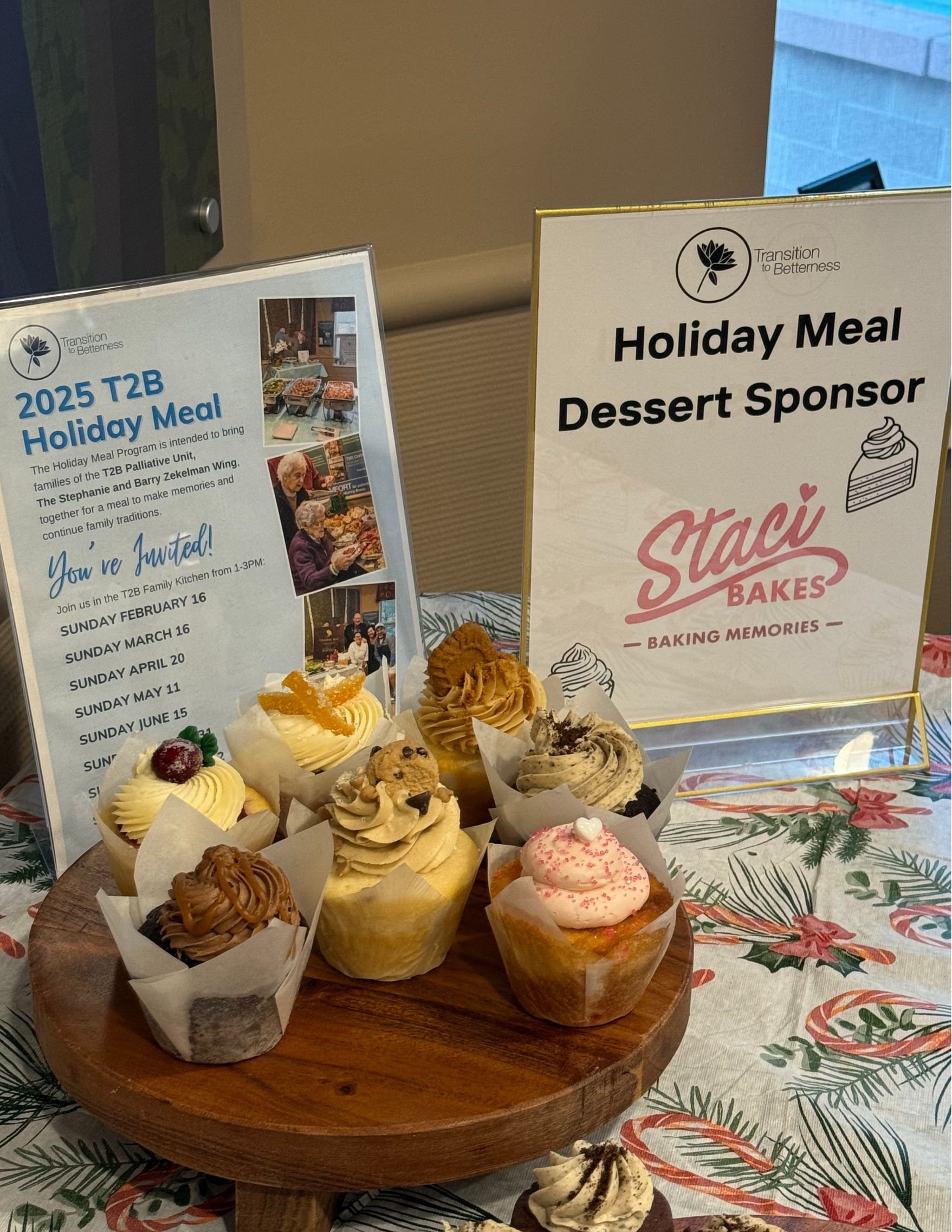 Assorted cupcakes on a wooden platter at a holiday event, with signs indicating a donation and sponsorship for a meal program.