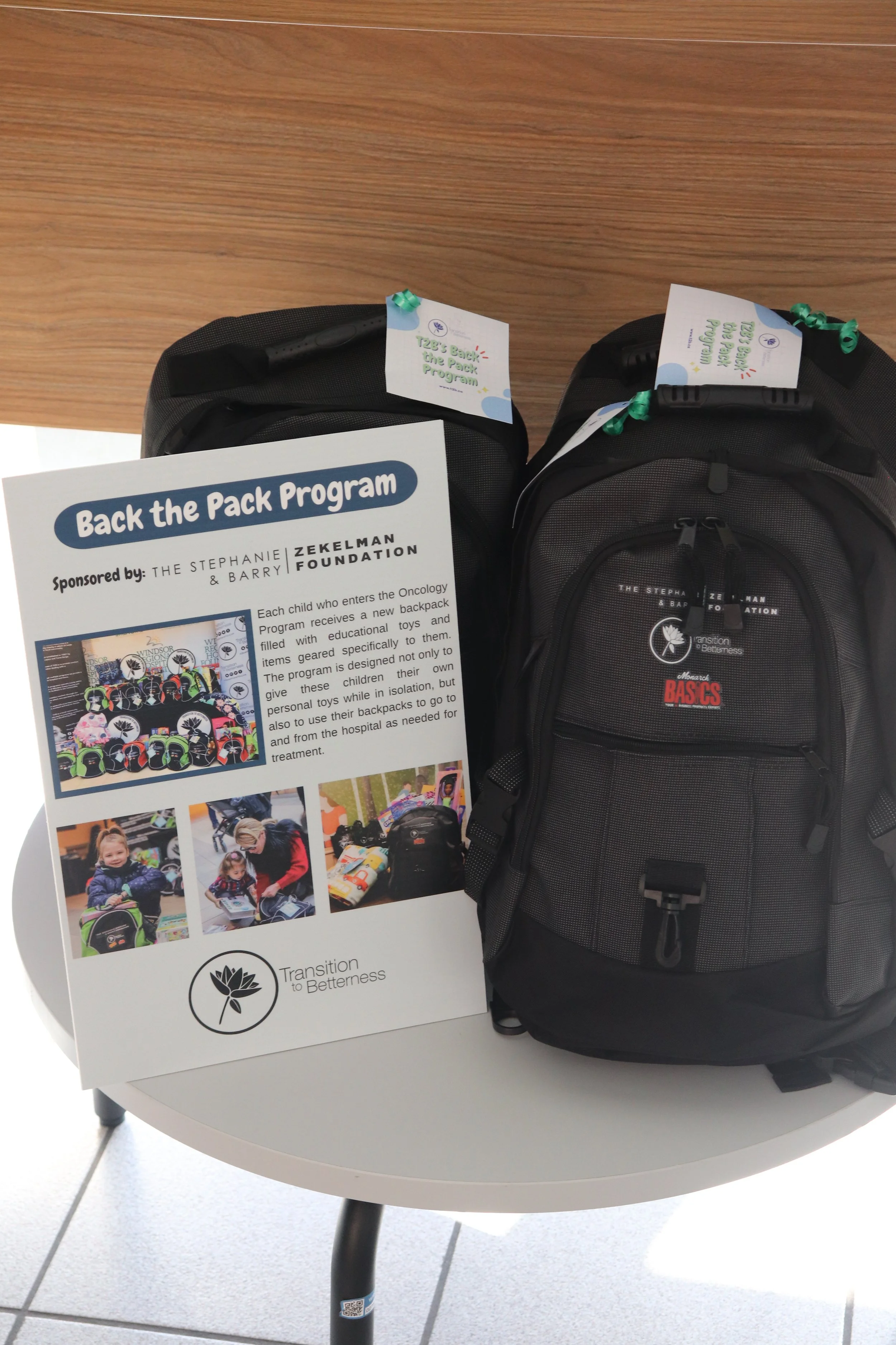 Two backpacks on a small table with a flyer promoting the Back the Pack Program by the Stephanie & Barry Zekelman Foundation, which provides backpacks filled with educational toys and items to children undergoing cancer treatment. The flyer includes images of children and backpacks.