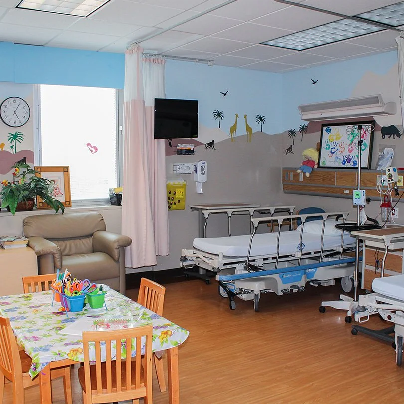 A pediatric hospital room with a medical bed, a wall mural of giraffes and other animals, a window with pink curtains, a beige sofa, a small table with children's art supplies, and medical equipment.