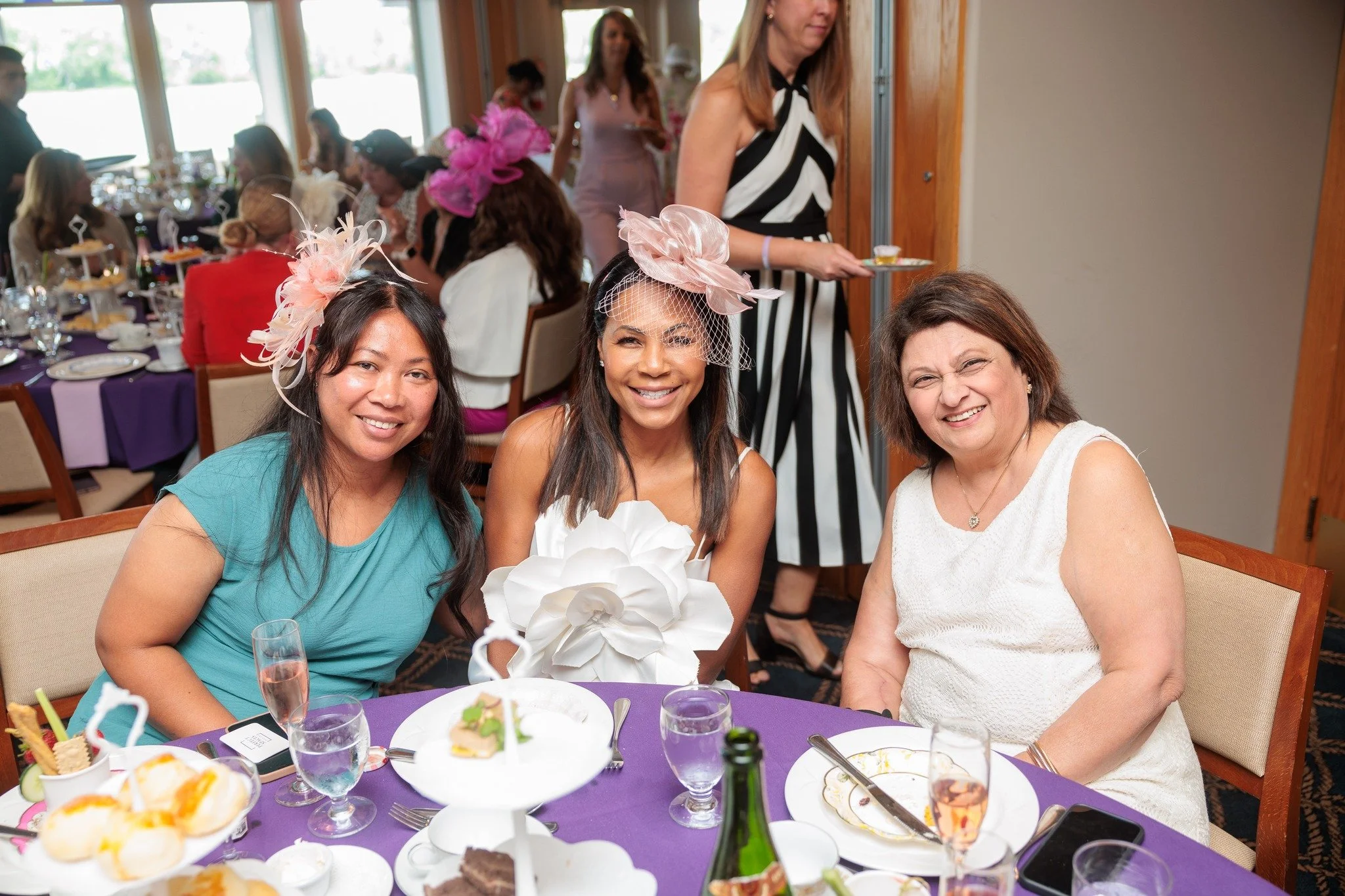 Three women sitting at a banquet table smiling at a celebration, with one holding a bouquet and everyone dressed in semi-formal attire, some wearing distinctive headpieces.