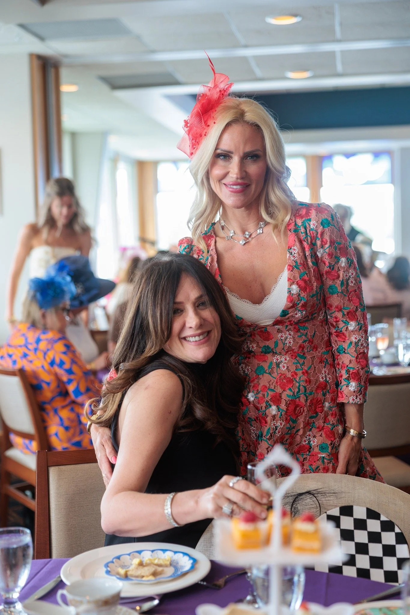 Two women at a celebration or party, one sitting and the other standing. The sitting woman has long dark hair and is wearing a black dress, smiling and holding a dessert. The standing woman has blonde hair, wearing a colorful floral dress with a pink