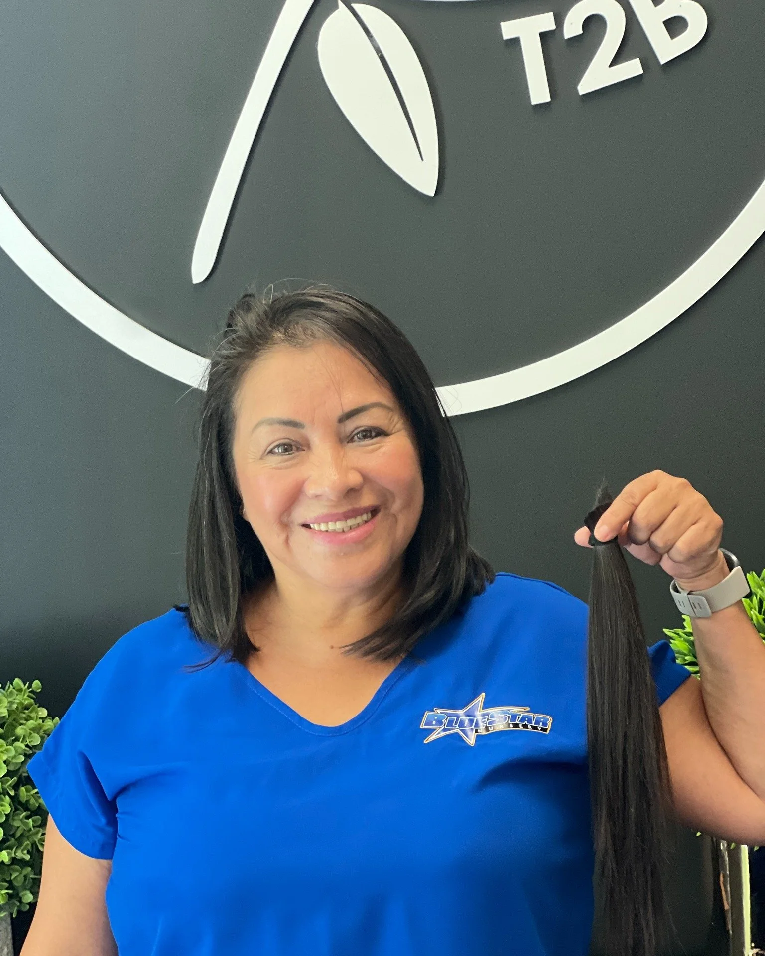 A woman smiling and holding a long, dark hair extension, standing in front of a black wall with a large clock and a logo for 'BlueStar' on her blue shirt.