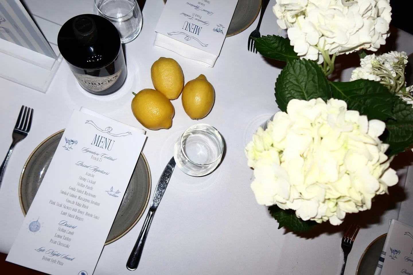 Elegant table setting with a menu, three lemons, a glass of water, a bottle of syrup, and white hydrangea flowers.