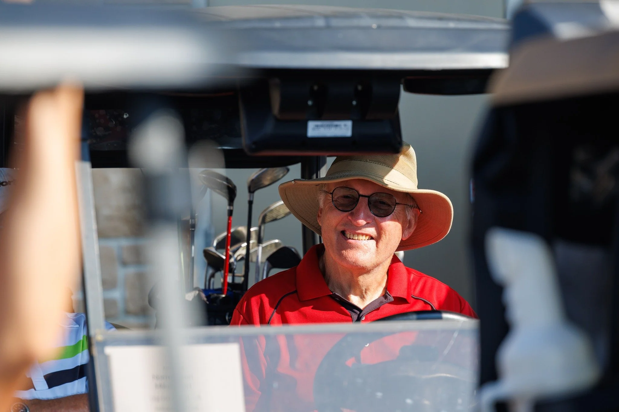 A smiling man with glasses wearing a wide-brimmed hat and red shirt, sitting in a golf cart on a golf course, with golf clubs behind him.