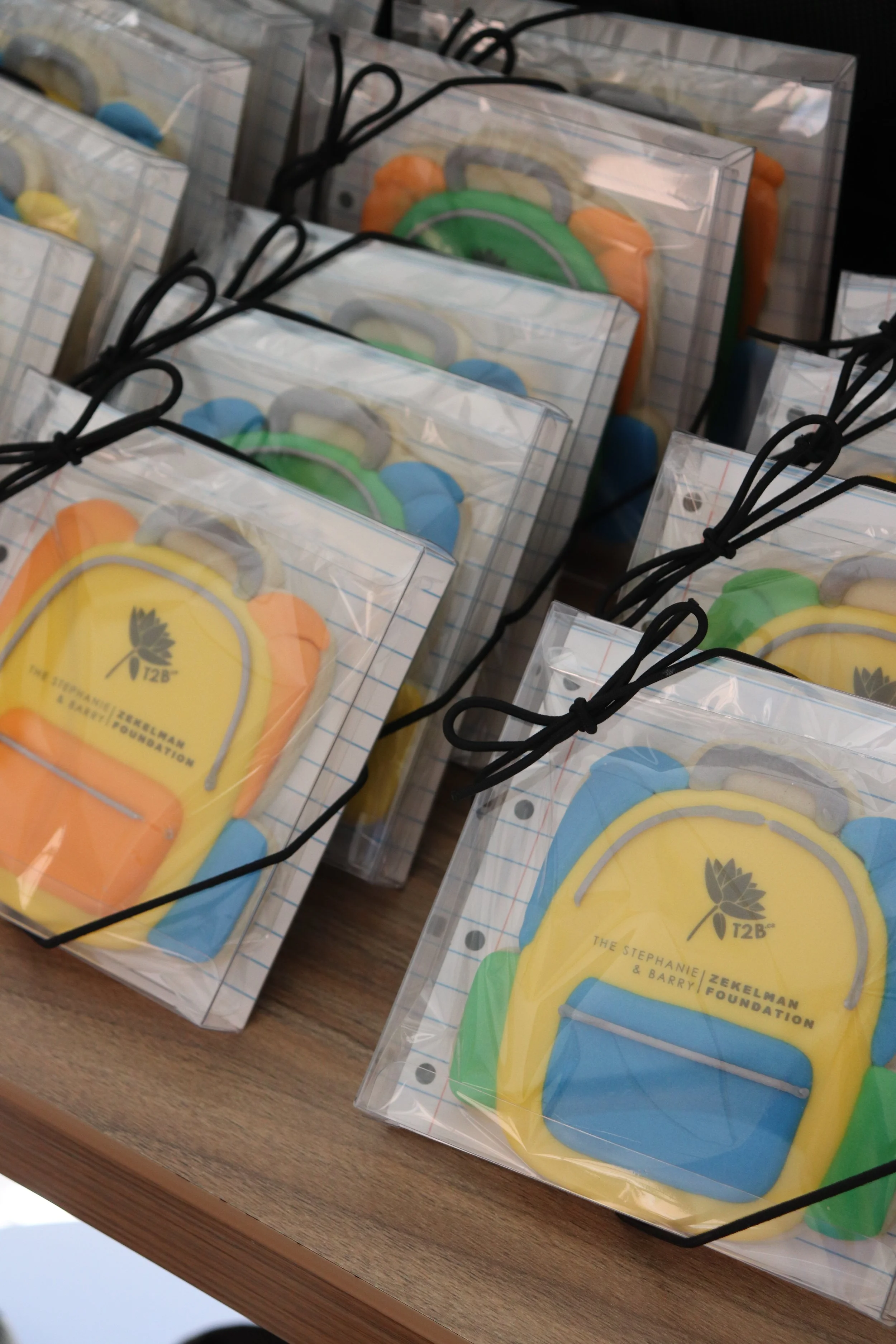 Colorful backpack-shaped cookies in clear plastic boxes with black ribbon ties, displayed on a wooden surface.