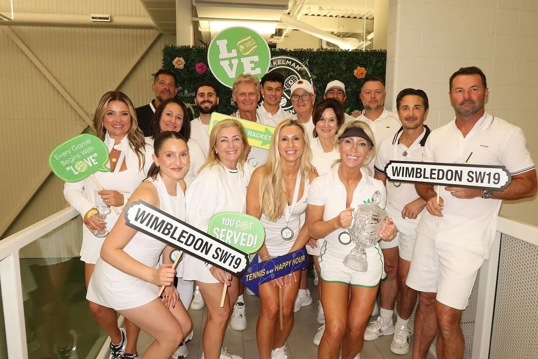 Group of people in white tennis attire celebrating at Wimbledon, holding signs with messages like "Wimbledon SW19", "You get served!", and "Every game begins with love"; some with trophies, with a green backdrop and flowers.