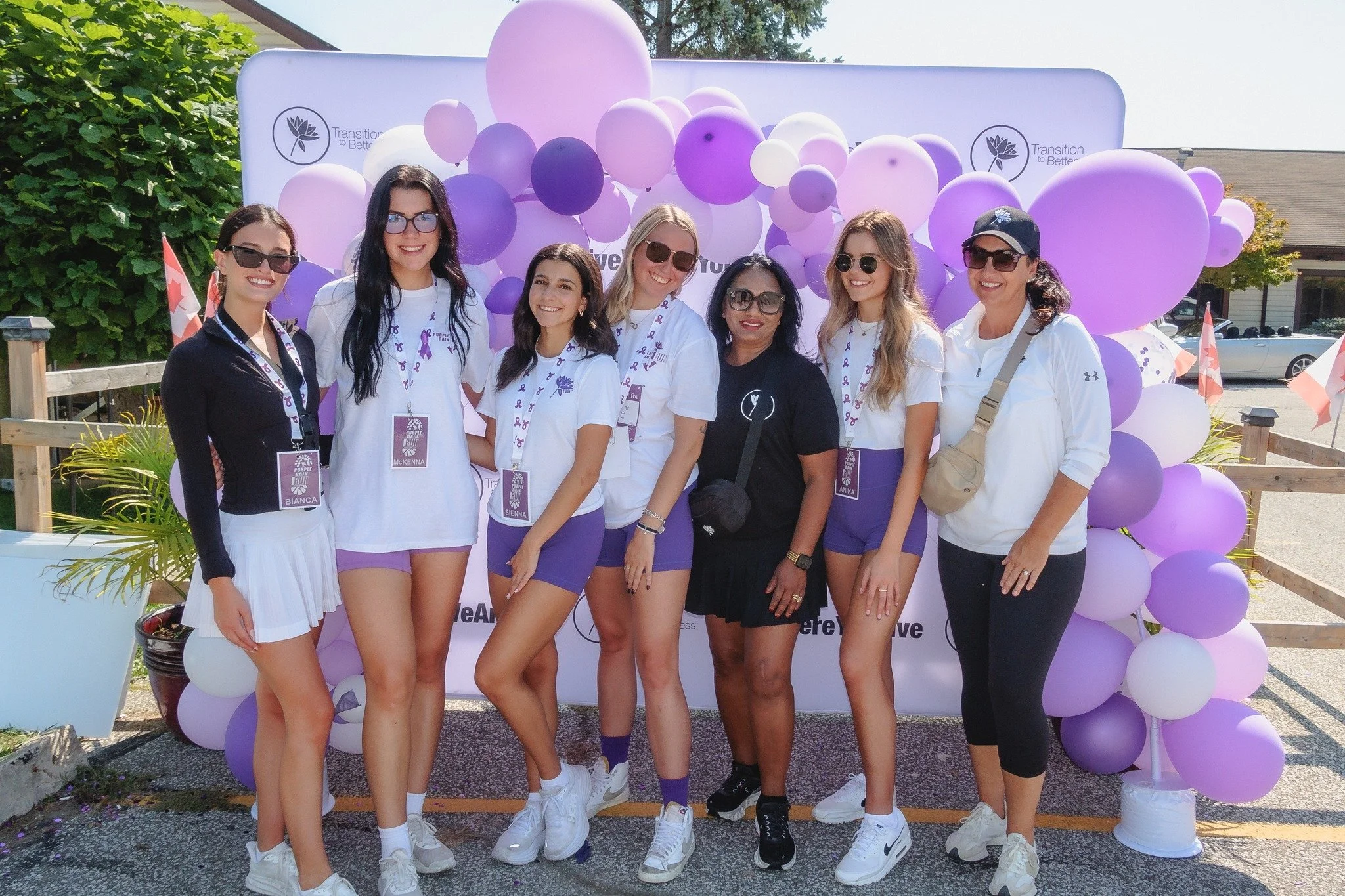 Group of eight women standing in front of purple and white balloons, posing for a photo. Most women are wearing white shirts, purple shorts or skirts, and sunglasses, with bibs displaying names and a logo. One woman wears a black shirt and skirt. They are outdoors, with a wooden fence, potted plants, and buildings in the background.