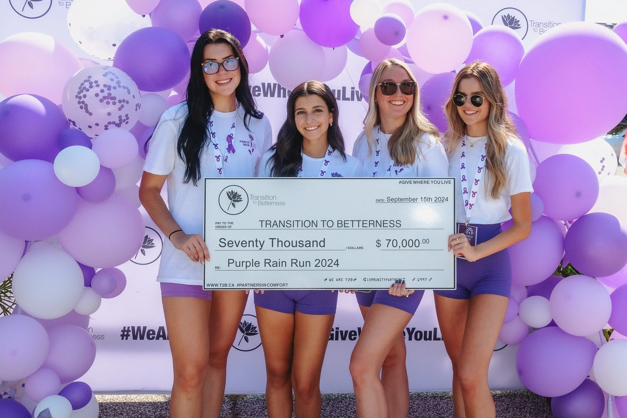 Four women standing in front of a purple balloon arch holding a large check for $70,000 made out to 'Transition to Betterness', dated September 15, 2024, participating in the Purple Rain Run 2024 event.