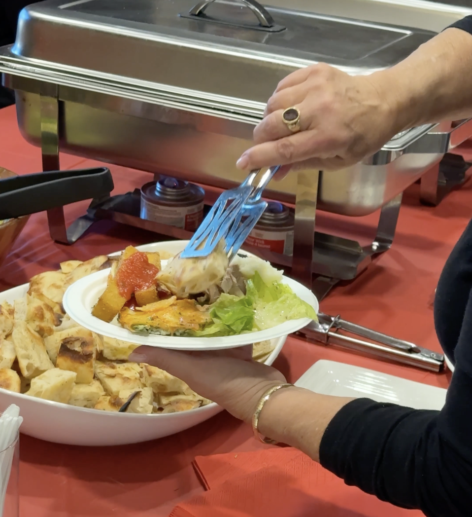 A person serves themselves food from a buffet, placing salad on a plate that already has some quesadillas. The buffet includes a chafing dish and bowls of food, with a red tablecloth underneath.