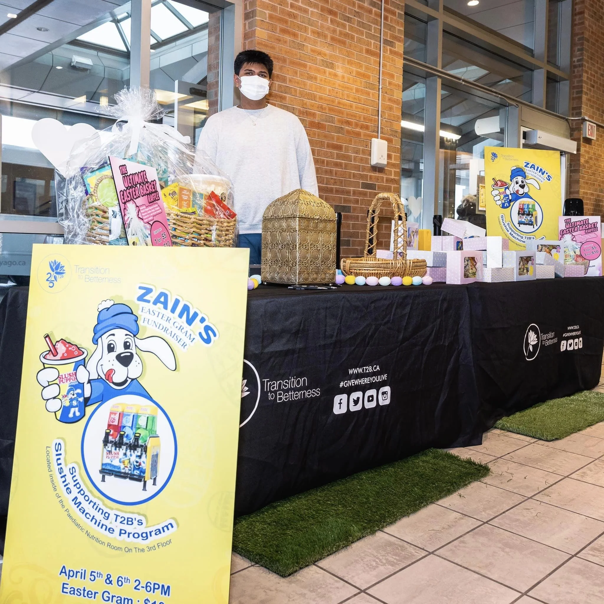 A person wearing a white face mask and a light gray sweatshirt standing behind a table with Easter-themed baskets and raffle prizes. The table has a black tablecloth with the logo and website of Zain's Easter Gram Fundraiser, and a yellow poster with