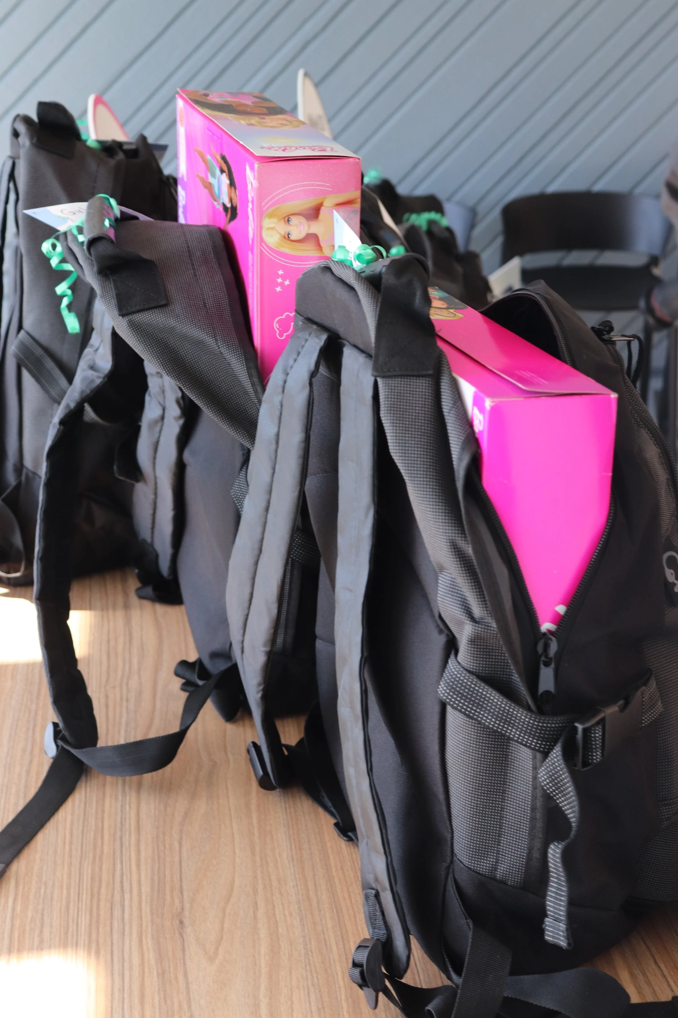 Three backpacks with pink boxes on top, placed on a wooden table against a gray wall.